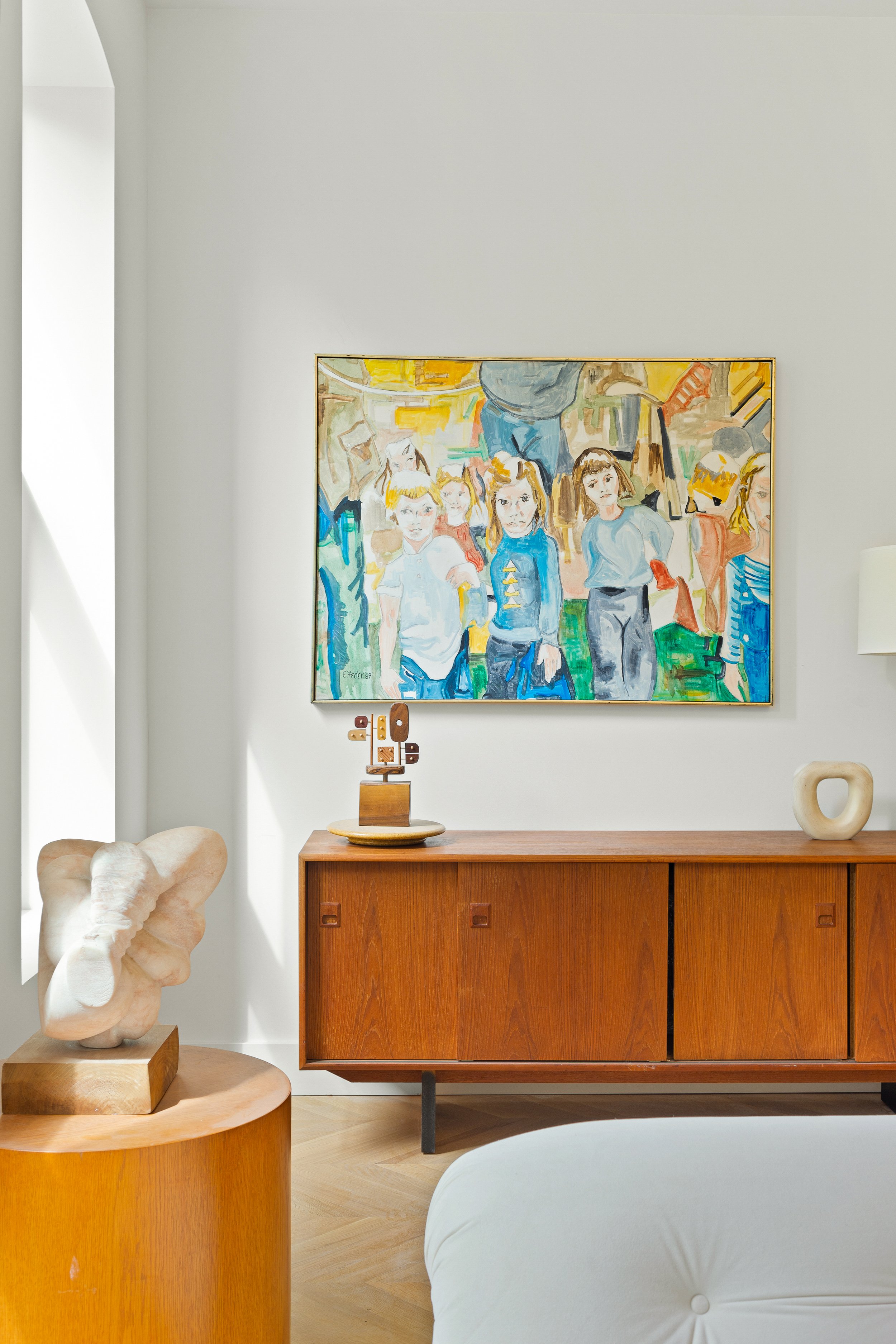Interior of a modern living space with a wooden sideboard, a colorful abstract painting of children hanging on a white wall, a wooden sculpture on a tray, a cream-colored sculpture, and part of a white sofa.
