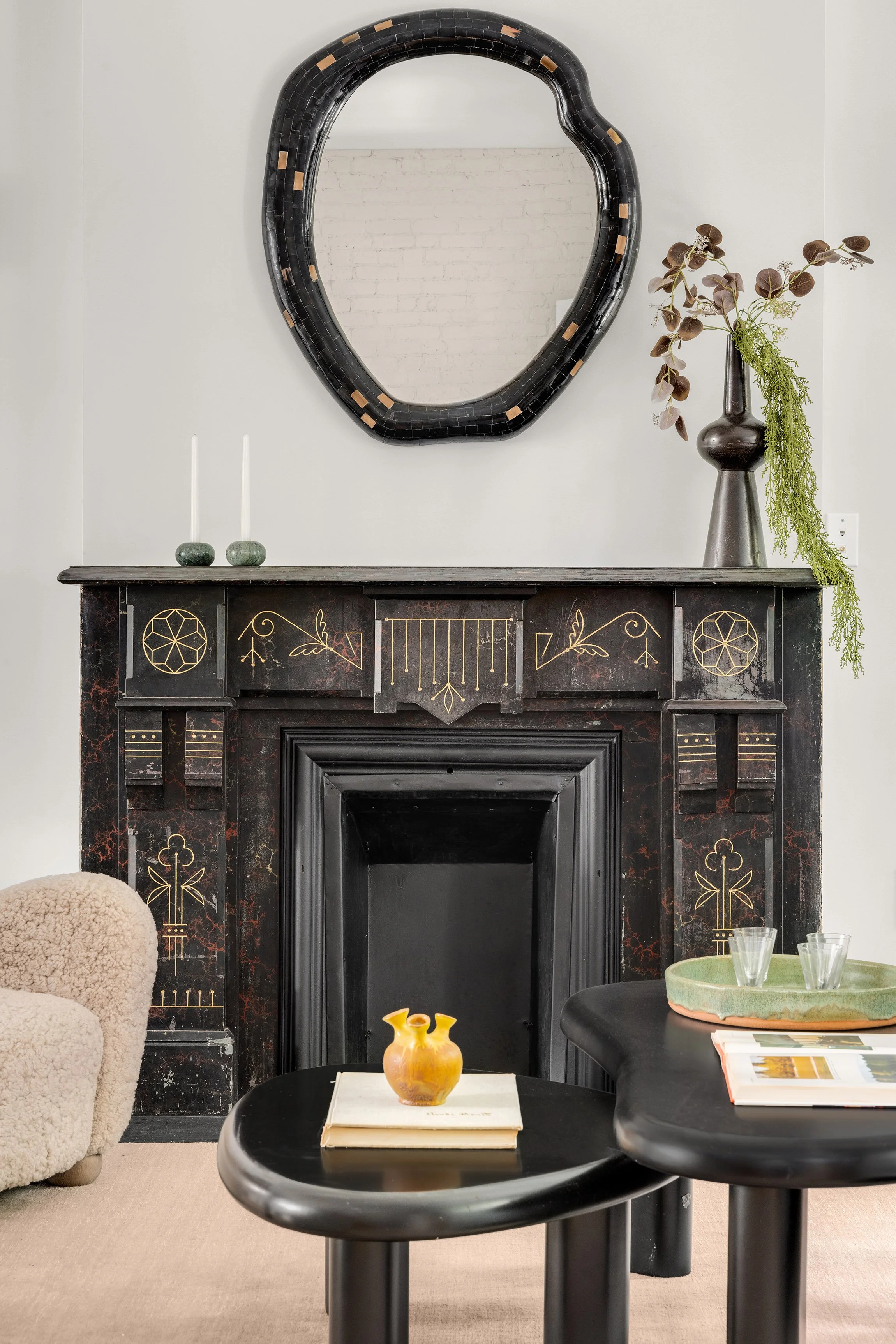 Interior living room with a black fireplace mantel decorated with gold details, a large irregularly shaped mirror above it, and a tall dark vase with greenery on the right. There is a cream-colored armchair on the left and black tables with a small vase, glasses, and books.