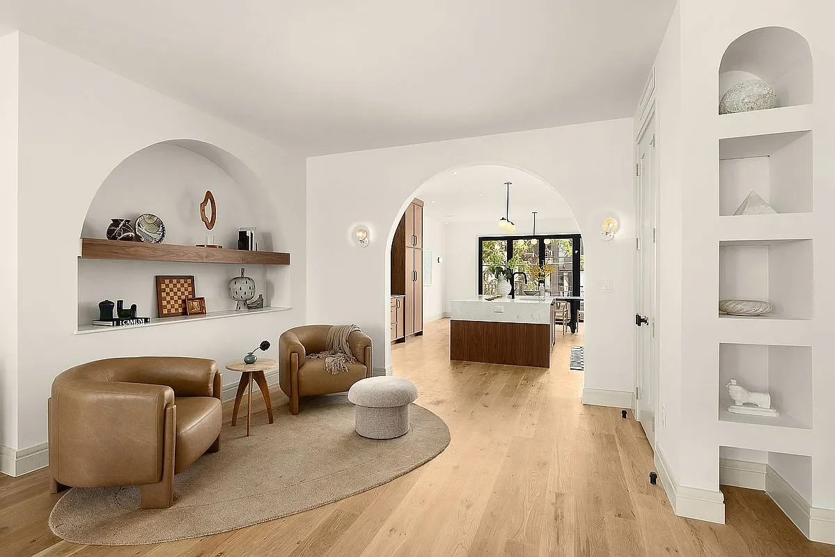 Open-concept living room with white walls, wooden flooring, two brown leather armchairs, a small round side table, and built-in shelves displaying decorative objects. Beyond, a kitchen with a large island, wooden cabinets, and black-framed glass doors that lead outside.