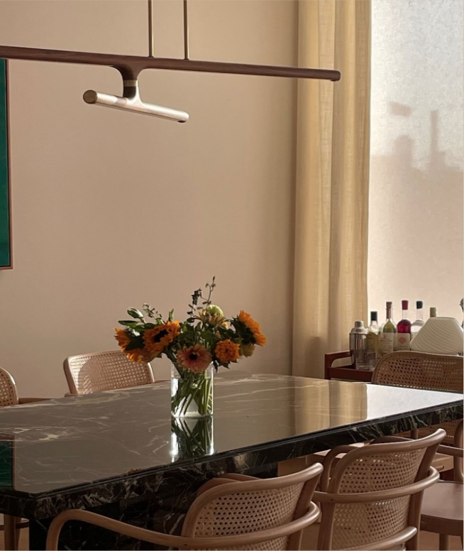 Decorative dining room table with a vase of sunflowers and other flowers, surrounded by wicker chairs, in a room with beige walls, a ceiling light fixture, and a window with beige curtains.