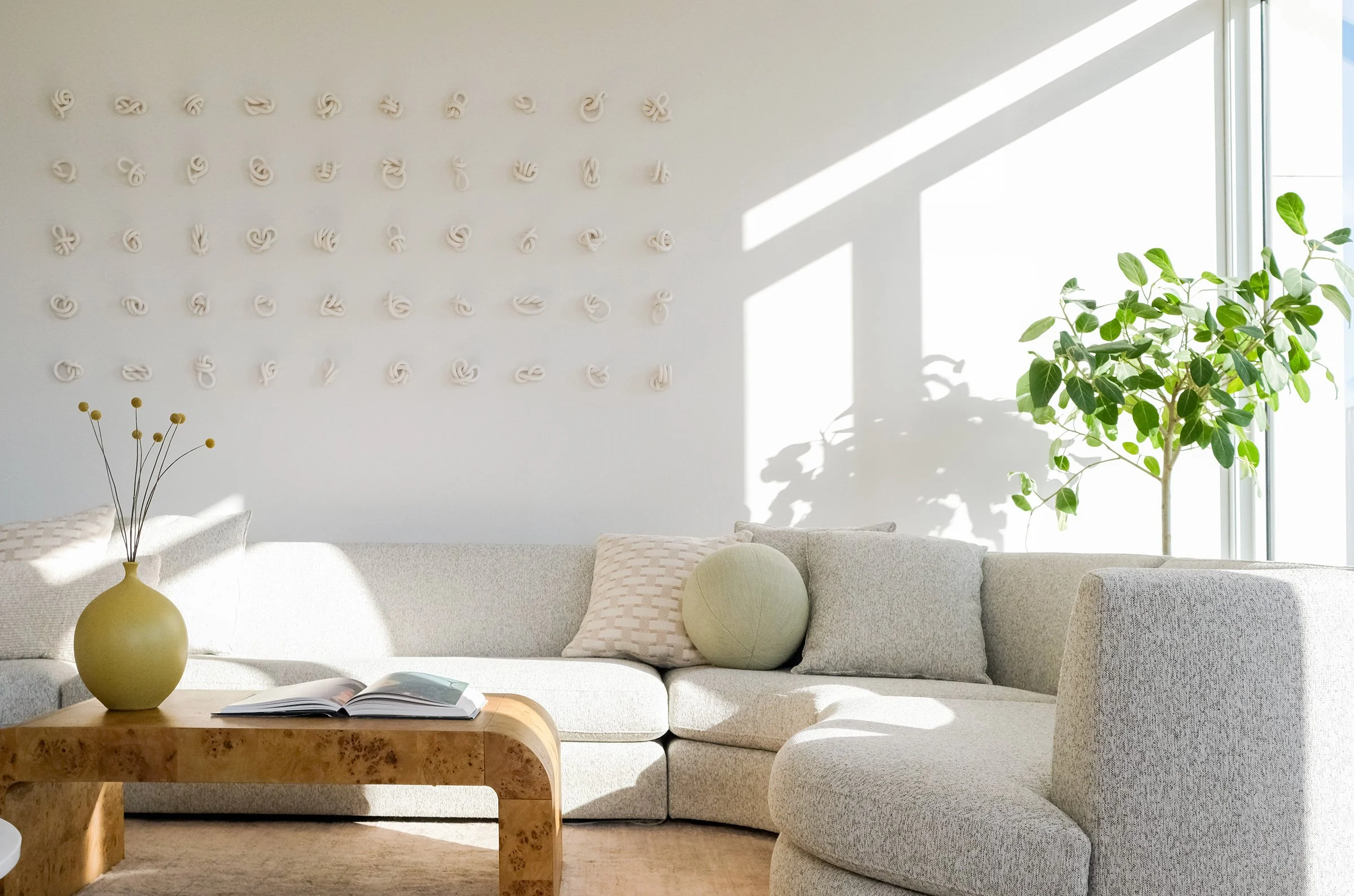 Bright, modern living room with white sofa, wooden coffee table, green plants, wall art, and sunlight streaming through large windows.