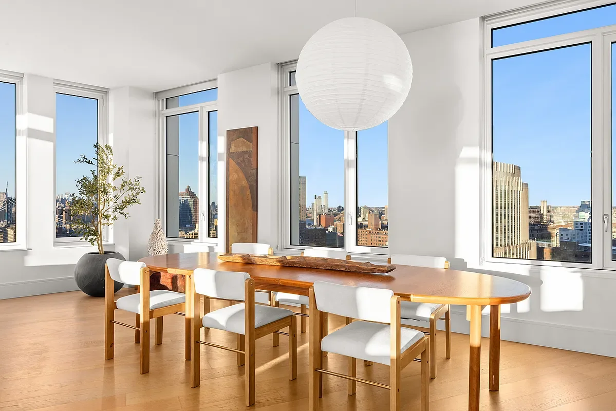 Bright dining room with large windows showing city skyline, wooden dining table with six white chairs, potted plant, abstract artwork, and large white paper lantern hanging from ceiling.
