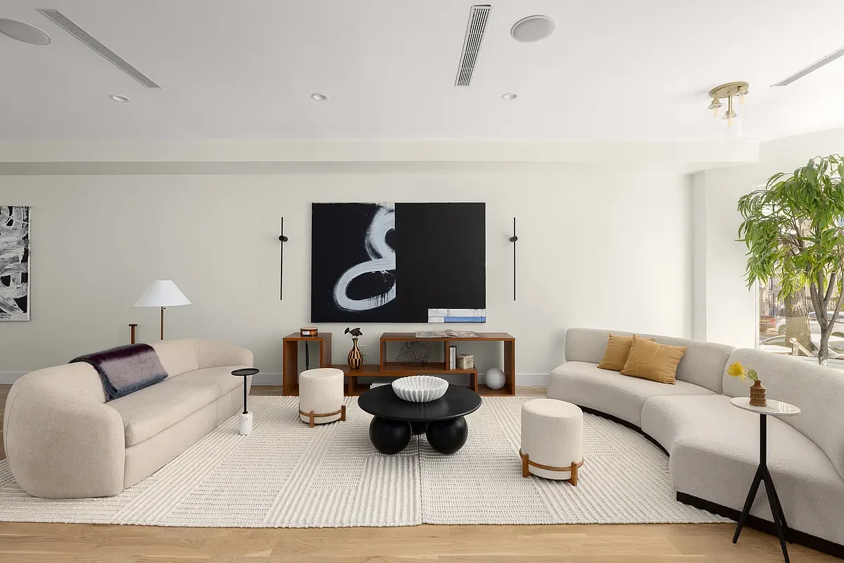 A modern living room with white walls, beige sofas, a black round coffee table, a white textured rug, and a flat-screen TV on the wall. There are decorative pillows, a floor lamp, and a potted plant near the window.