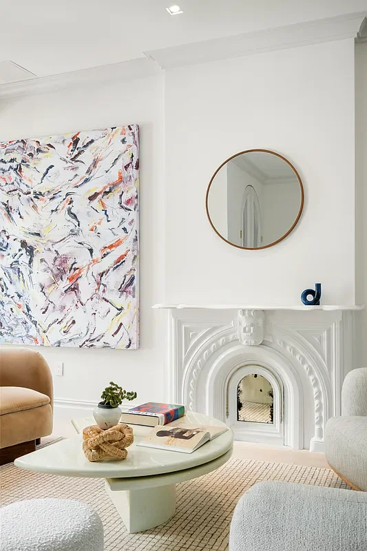 Living room with white walls and fireplace, round mirror above fireplace, colorful abstract painting on wall, beige and gray furniture, and a small coffee table with books and decorative items.