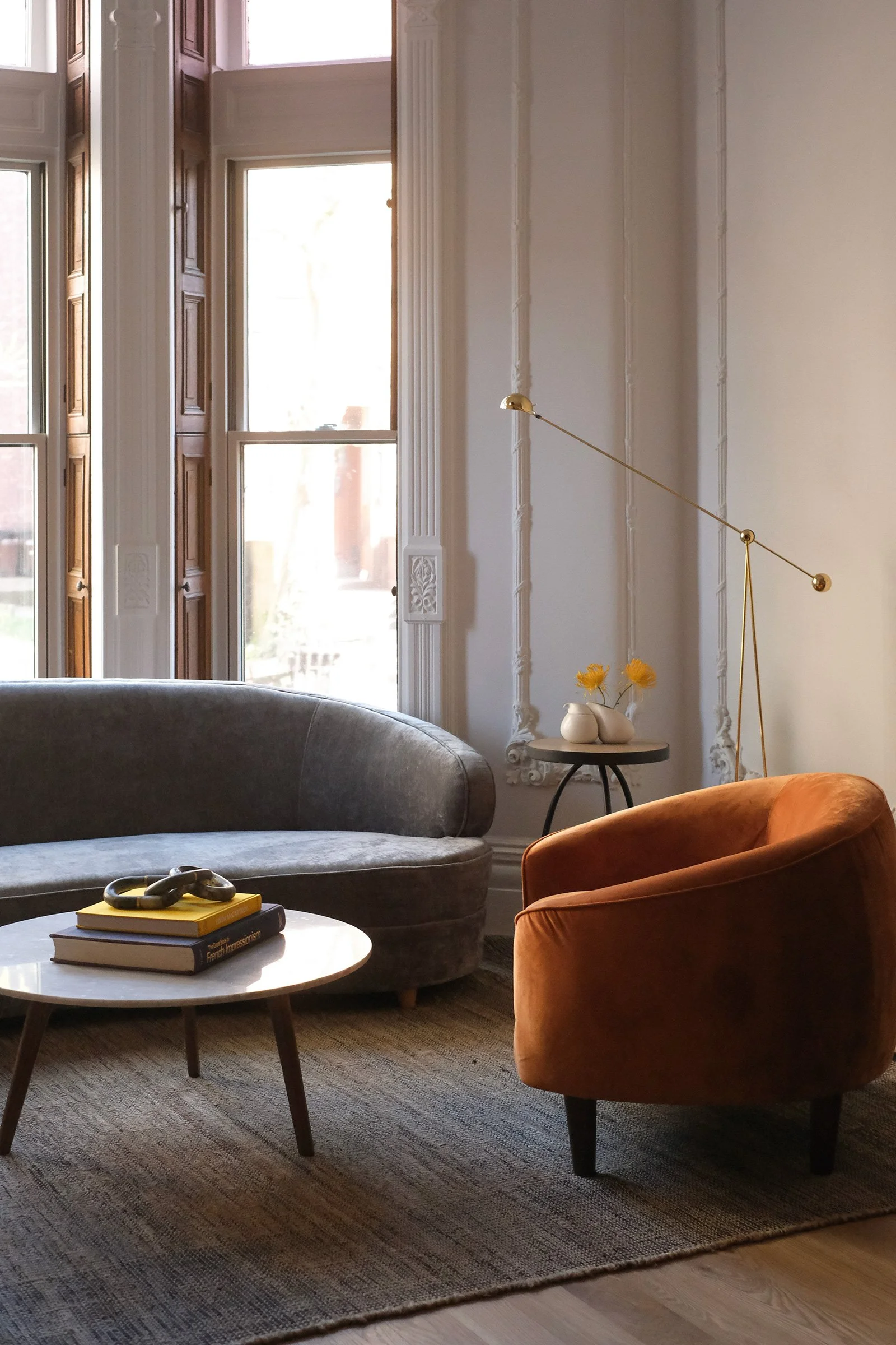 Living room with large windows, a grey velvet sofa, an orange armchair, a round coffee table with books and a decorative object, a small side table with vases and flowers, and a modern gold wall lamp.