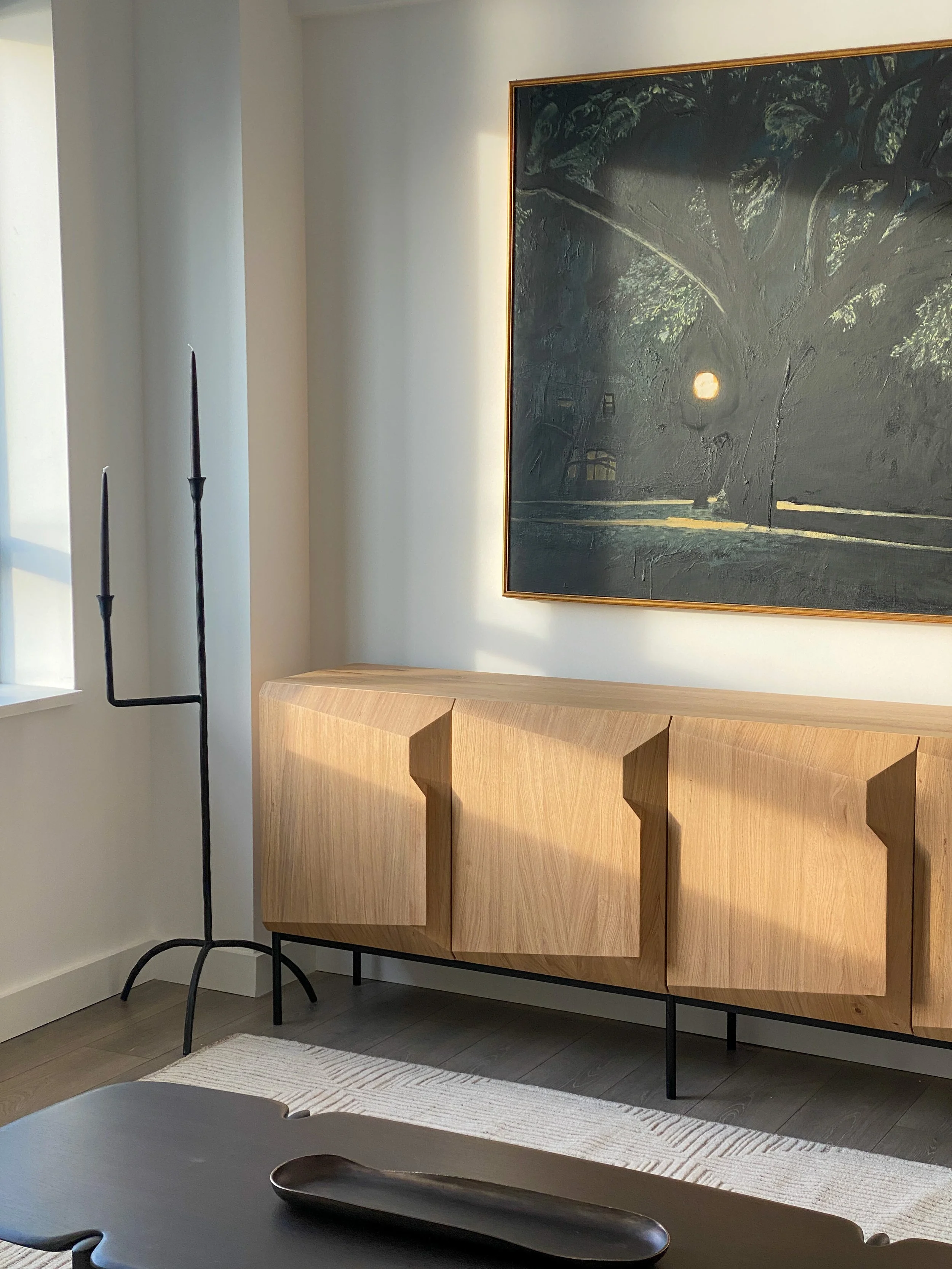 A modern living room corner featuring a wooden sideboard with a geometric design and black metal legs, a large framed painting of a tree with the moon, a black floor lamp with three thin candle-like holders, a window with sunlight streaming in, and a small portion of a dark coffee table with a black tray.