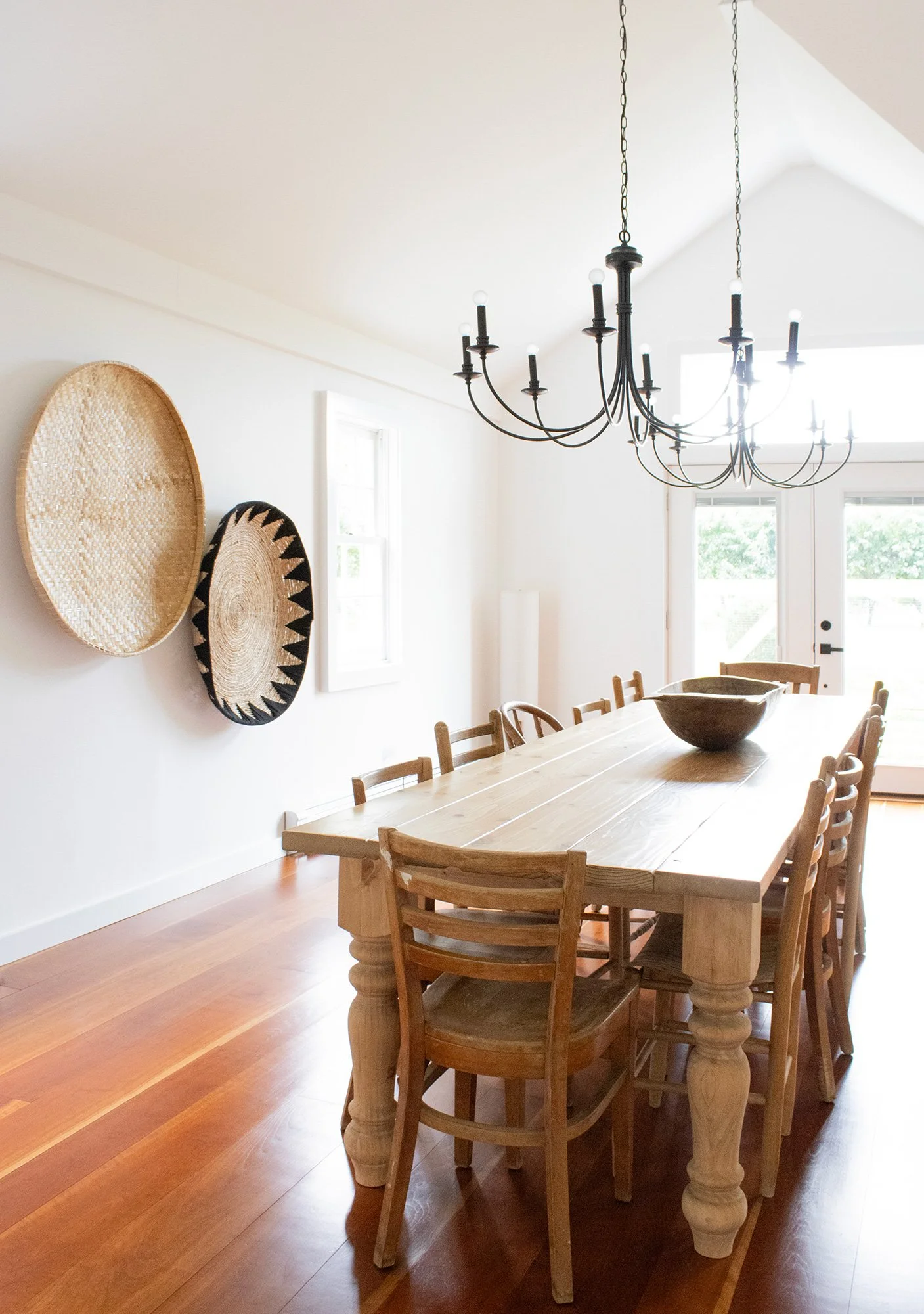 A dining room with a wooden table, eight chairs, a black chandelier, and woven baskets on the wall. There is a door and windows in the background.