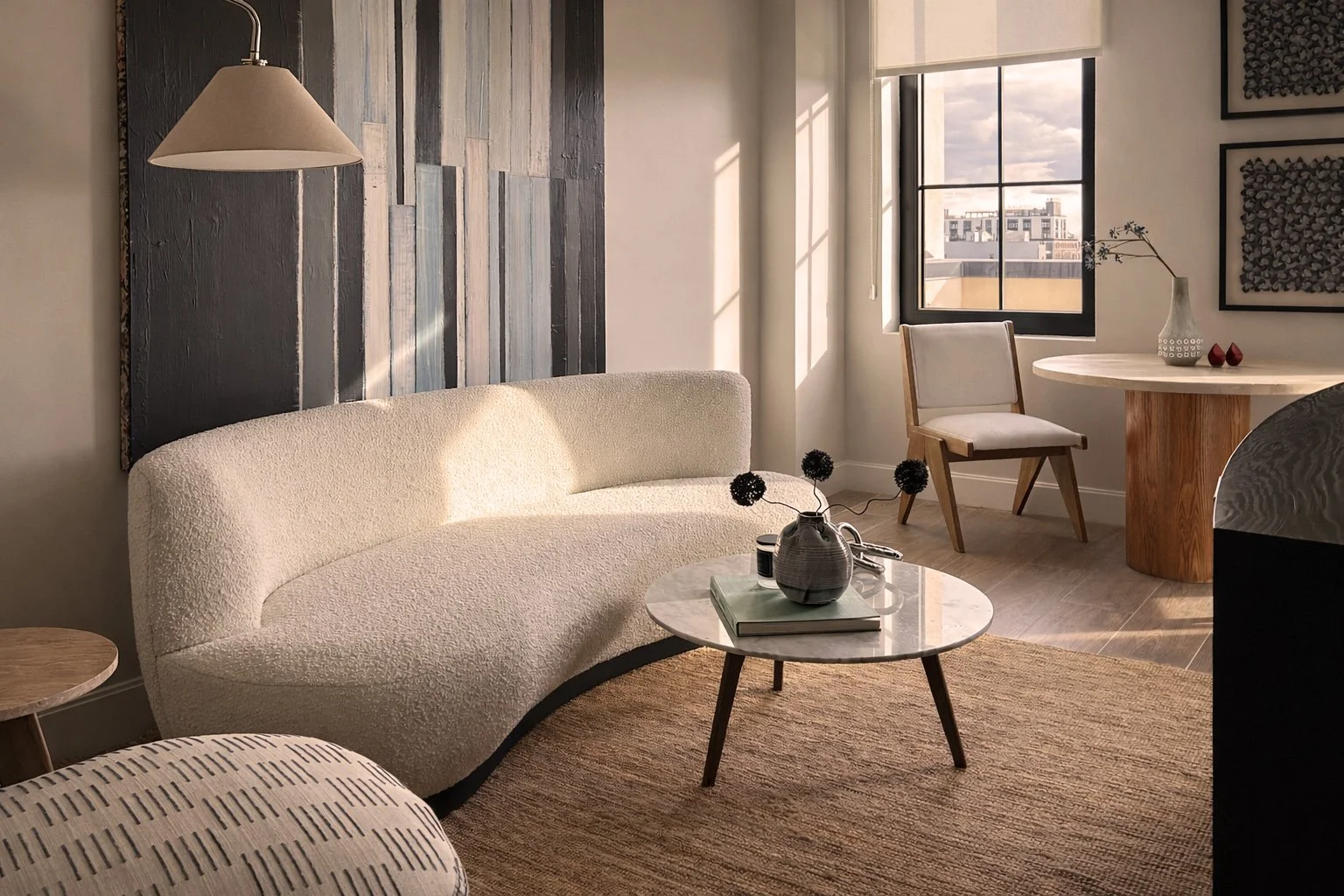 Bright living room with a beige textured sofa, round marble coffee table with black decorative flowers, wooden side table, armchair near window, and artwork on the walls, illuminated by sunlight through large window.
