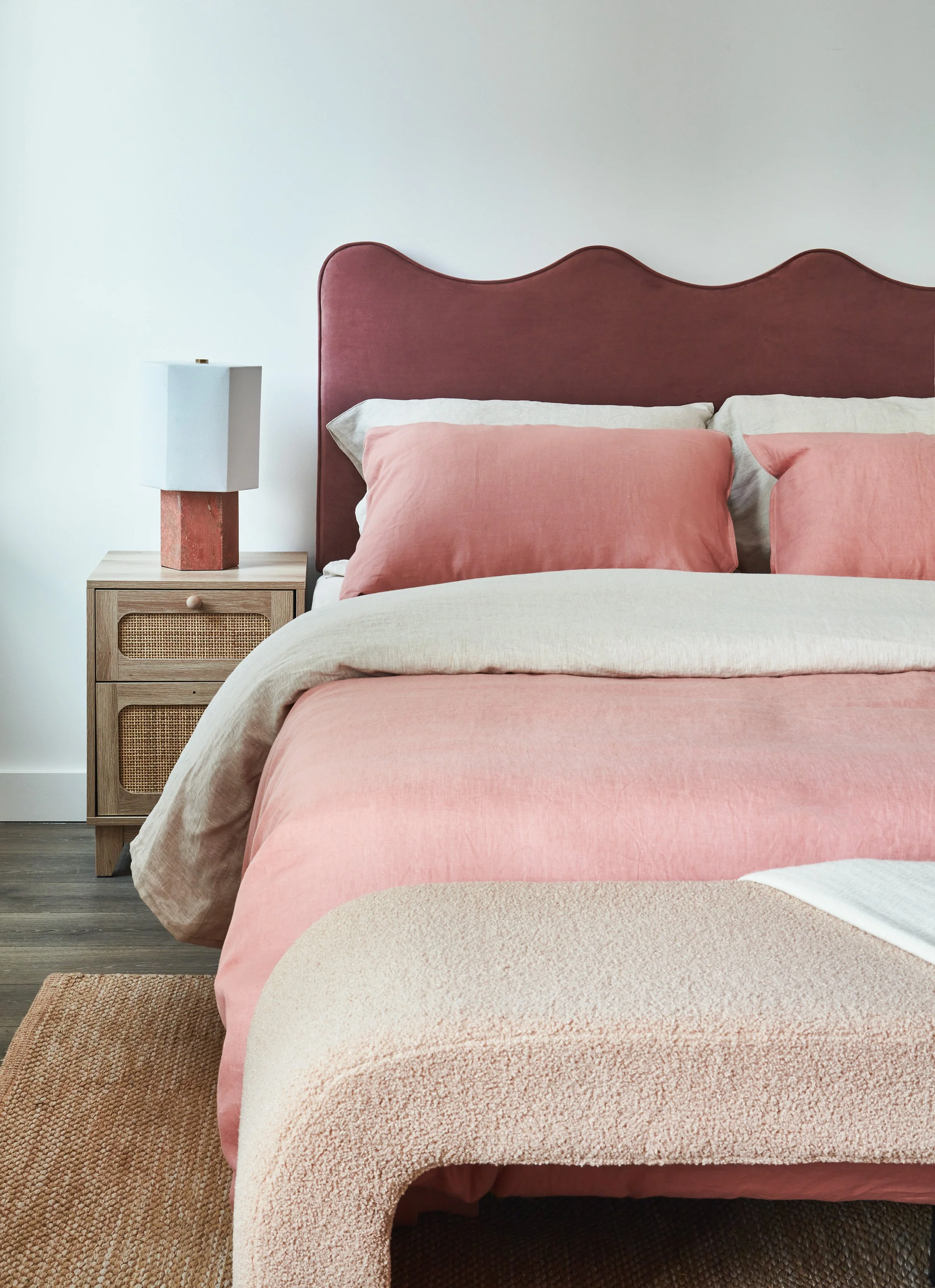 A neatly made bed with a pink velvet headboard, beige bedding, and pink pillows in a modern bedroom. Next to the bed is a wooden nightstand with a lamp and a white lampshade.