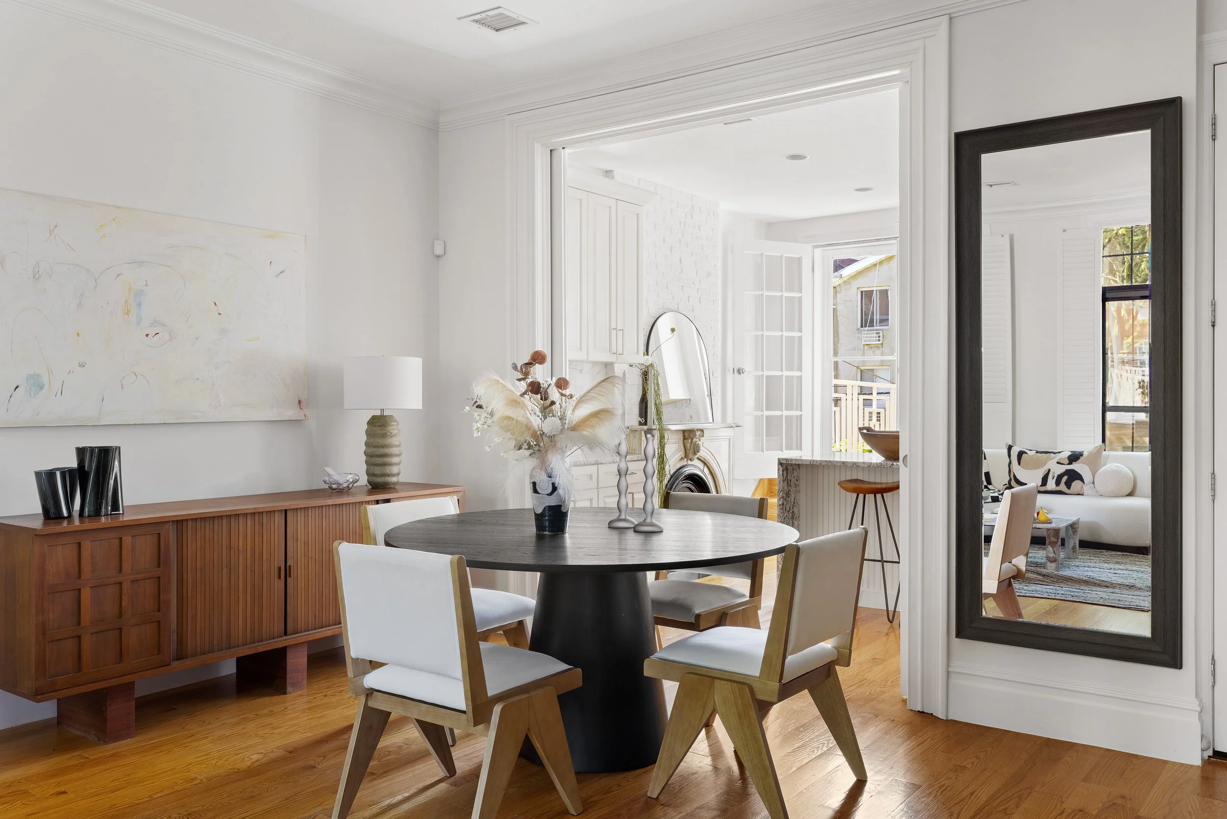 Modern dining room with black round table, four white chairs, wooden sideboard, large mirror, white walls, hardwood flooring, and adjacent living room with black and white decor.