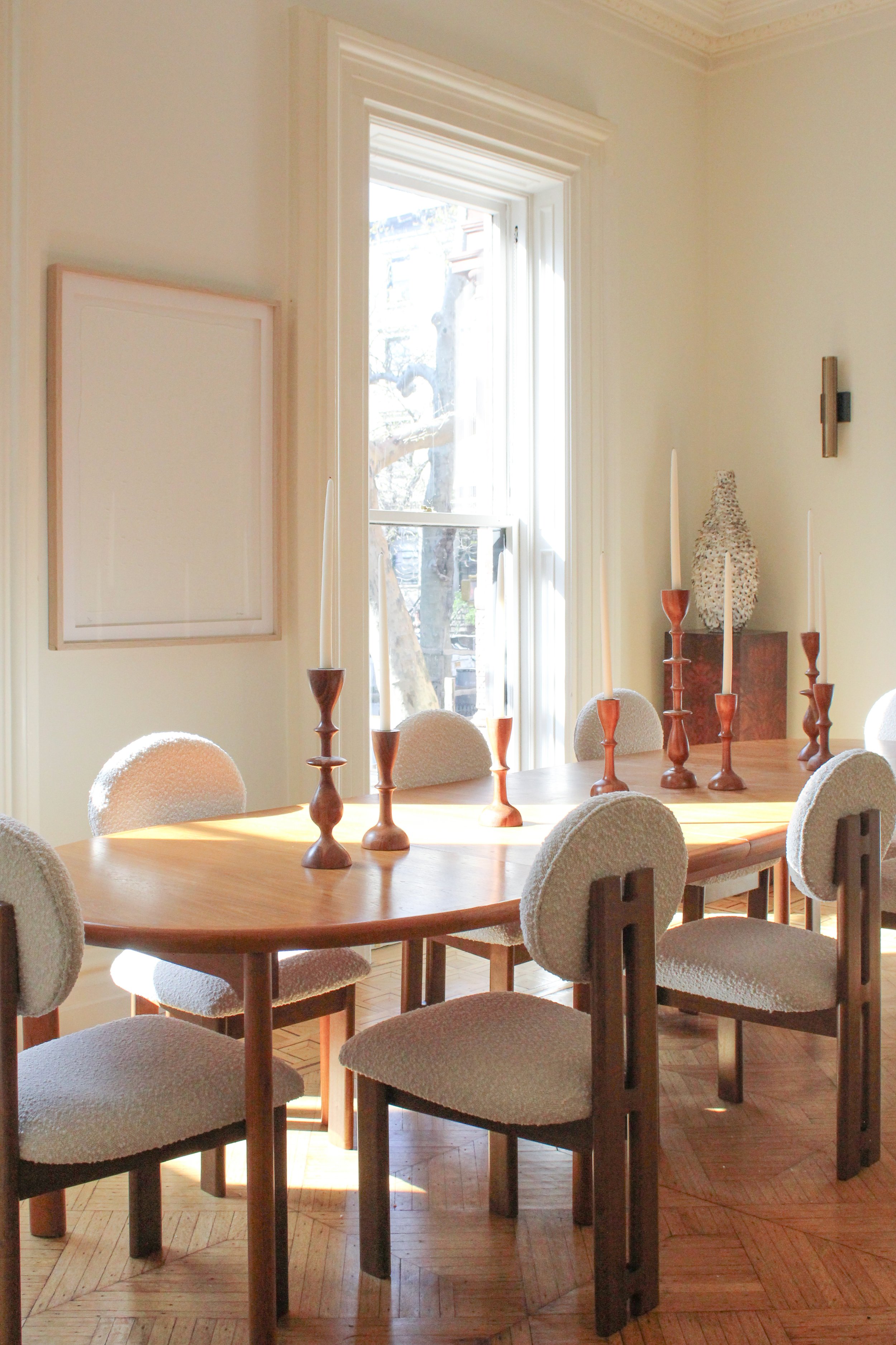 A dining room with a wooden table, six beige upholstered chairs, and seven tall wooden candlesticks with white candles. Sunlight streams through a large window, illuminating the room. There is a decorative vase on a side table in the corner and framed artwork on the wall.