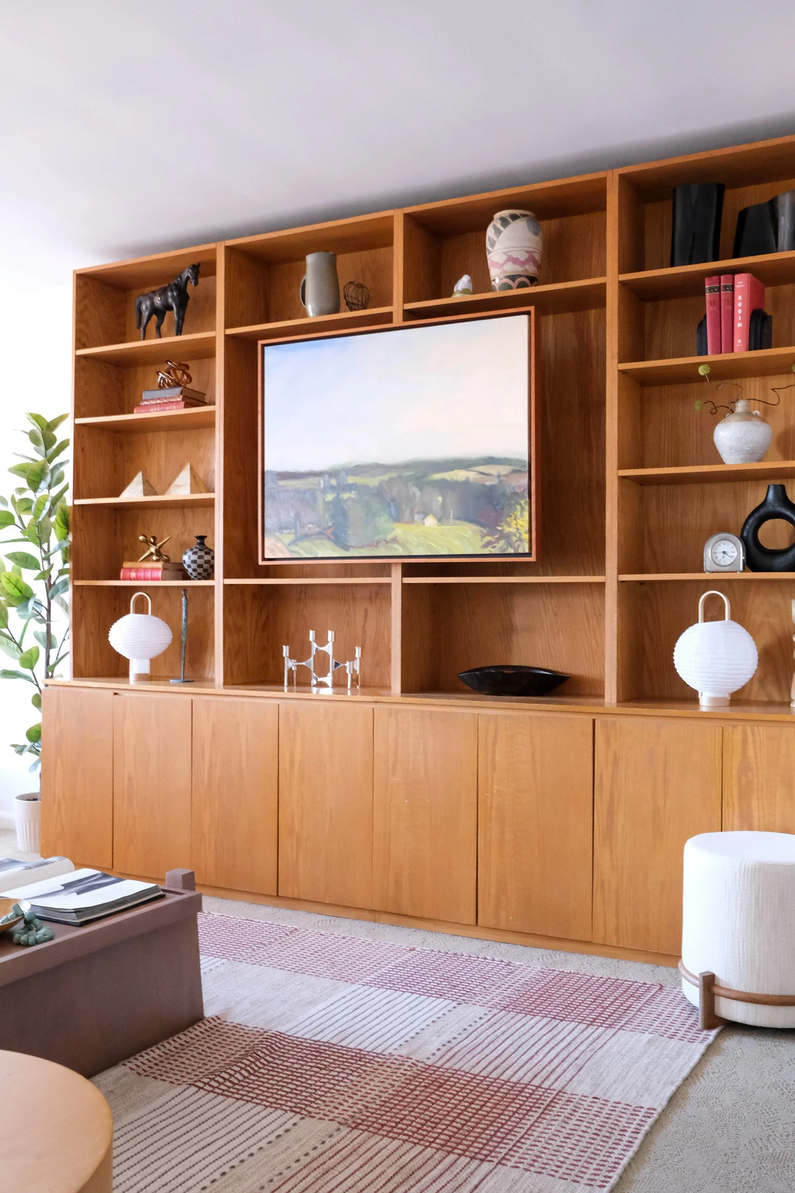 A wooden bookshelf with decorative items and a landscape painting in the center, located in a living room with a rug and a potted plant.
