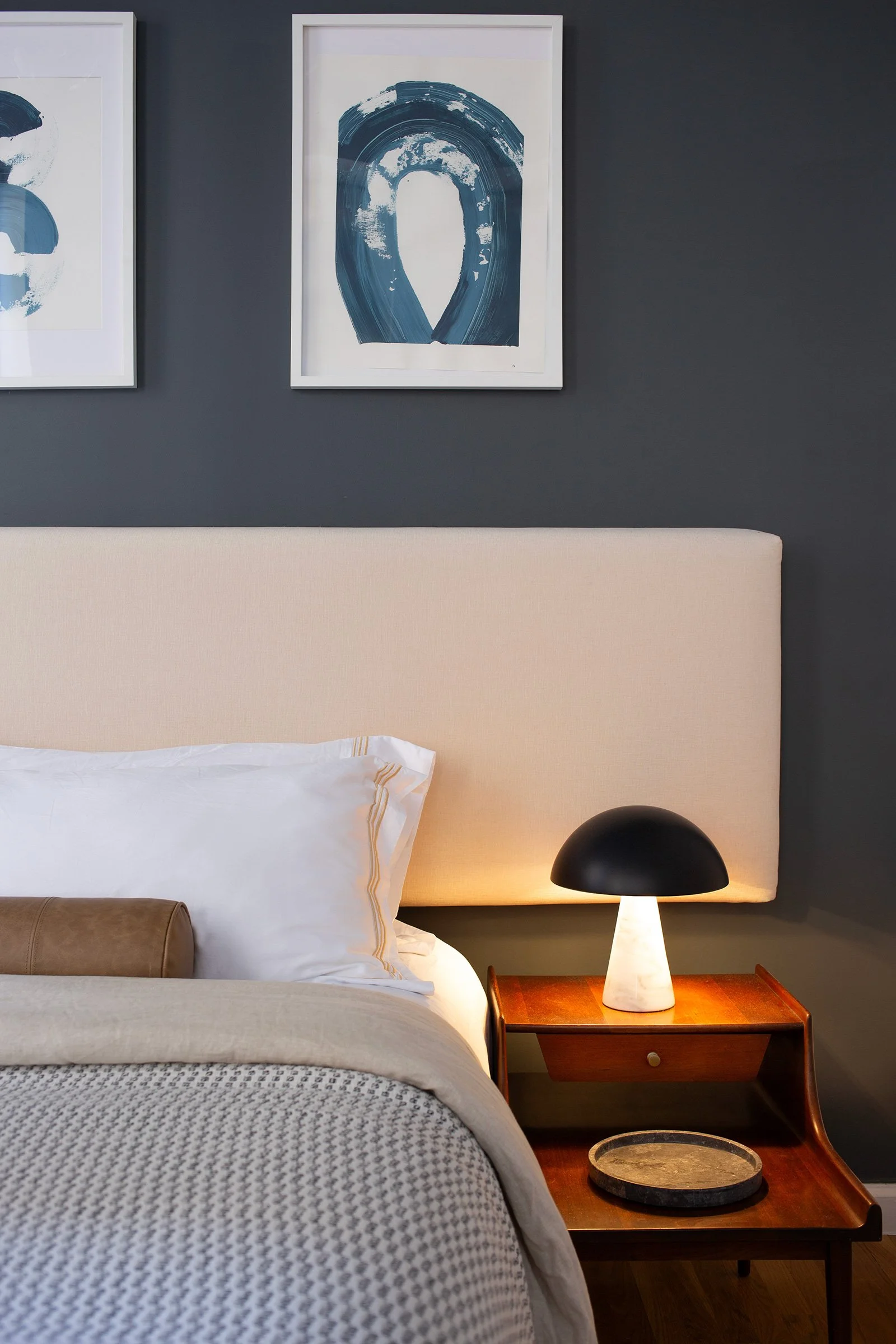 A bedroom with a bed, pillows, and a nightstand featuring a black and white table lamp. Abstract blue artwork hangs on a dark gray wall above the bed.