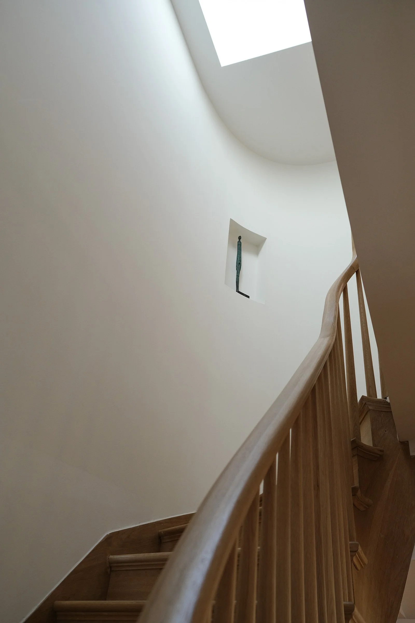 View of a spiral staircase with a wooden handrail and stairs, leading up to a bright skylight and a small alcove with a sculpture inside.