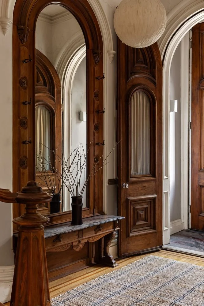 An interior hallway with a curved wooden door and matching wooden mirror frame, a white pendant spherical light, a vase with bare branches on a marble-topped wooden console, and a woven area rug on the hardwood floor.
