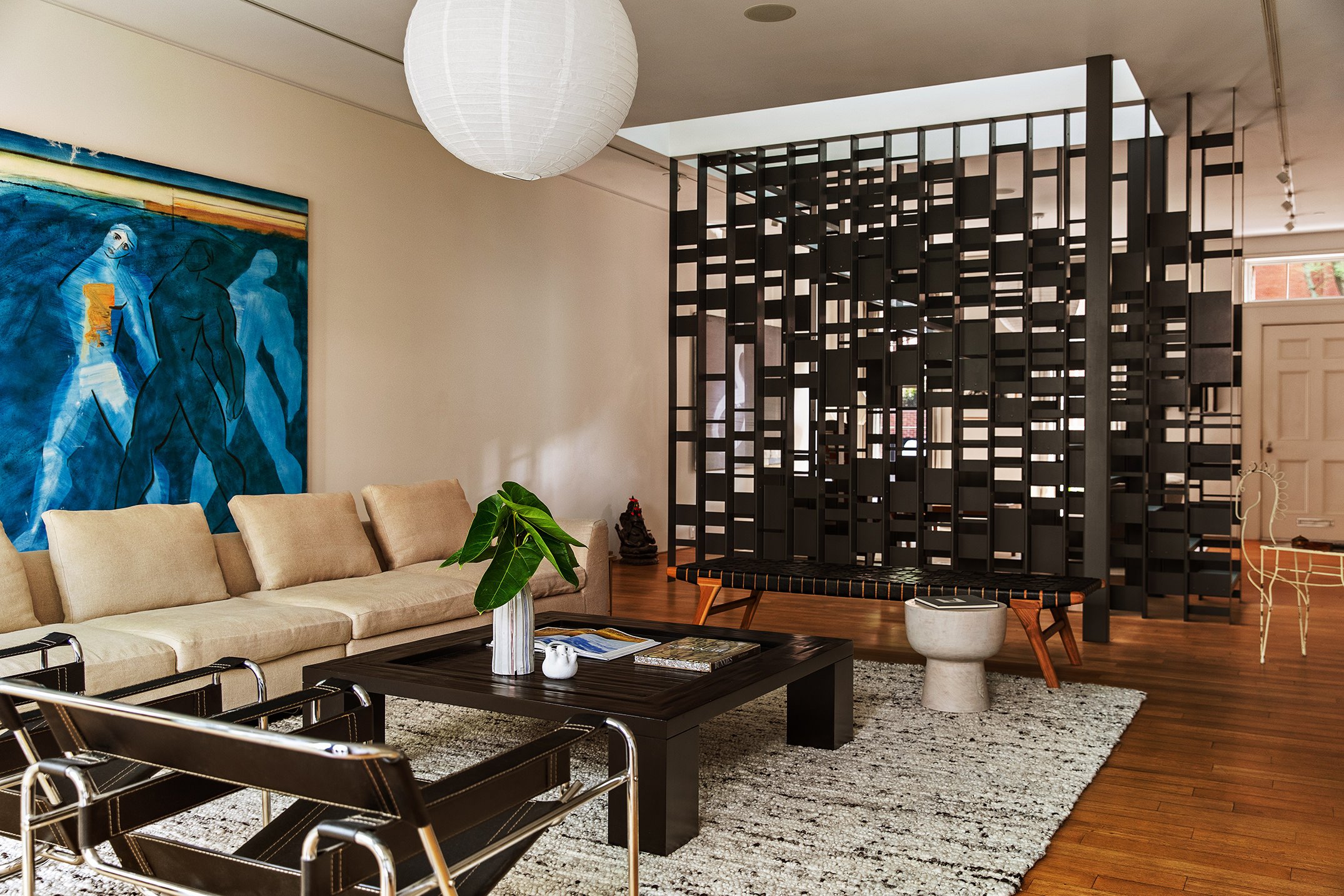 Living room with beige sofa, black coffee table with books, green potted plant, blue abstract painting, wooden stool, black wooden room divider, white door, and decorative iron chair in the background.