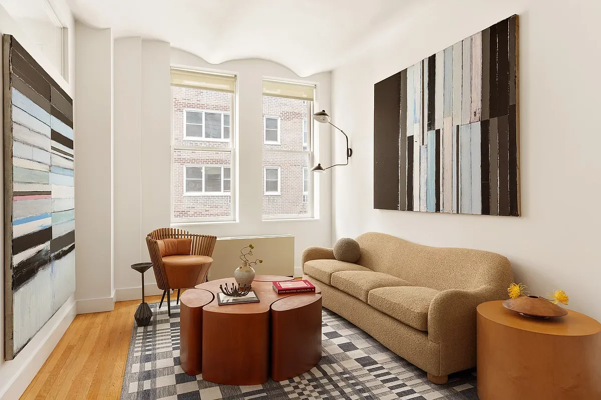 A modern living room with two large abstract paintings on white walls. The room features a beige sofa, a wooden coffee table, a round side table with a wooden tray and yellow flowers, and a tan armchair. There are two windows with a view of a brick building outside, and a black wall-mounted lamp.