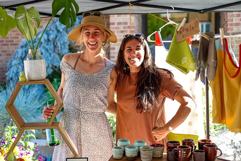 Two women smiling at a craft fair booth with plants and pottery