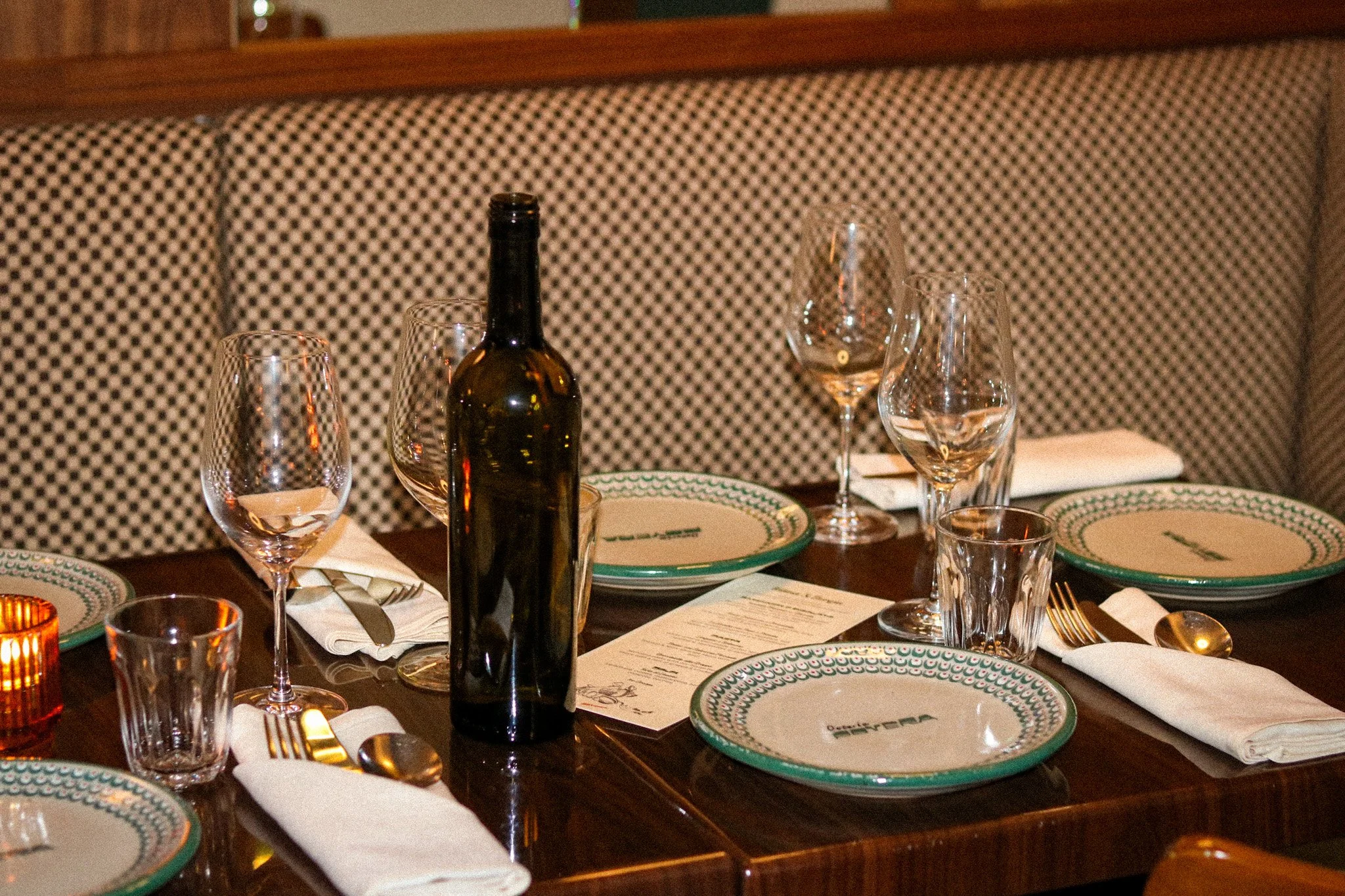 A restaurant table set with a patterned plate, a clear glass, a fork, a spoon, and a napkin with cutlery, with a padded booth seat in the background.