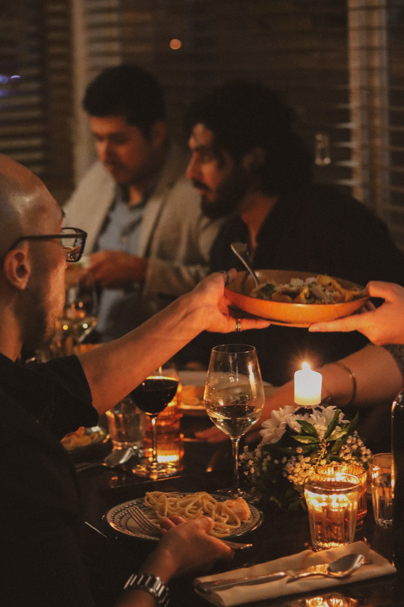 A plate of spaghetti with ground meat sauce on a dark wooden table, with a hand lifting a forkful of pasta. There are cherry tomatoes, a halved lime, and a glass of red beverage in the background.