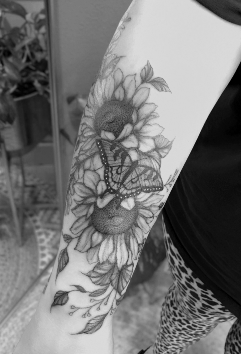Tattoo of a sunflower with a butterfly on the arm of a person wearing a black shirt and leopard print pants, with green plants in the background.