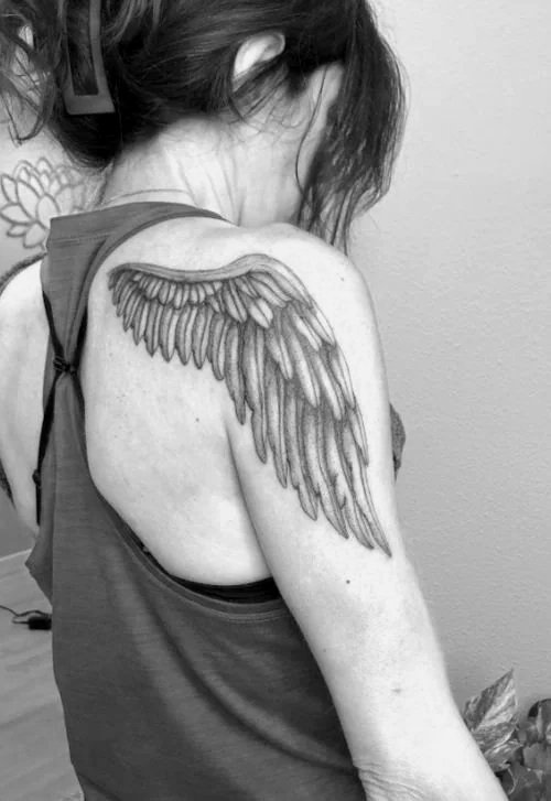 A woman with brown hair wearing a sleeveless shirt shows a large tattoo of angel wings on her left shoulder and upper arm.