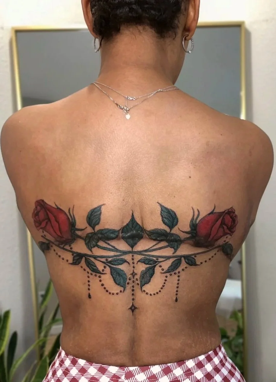 Back view of a woman with a floral tattoo of red roses and dark green leaves across her upper back, accented with black dots and decorative elements.