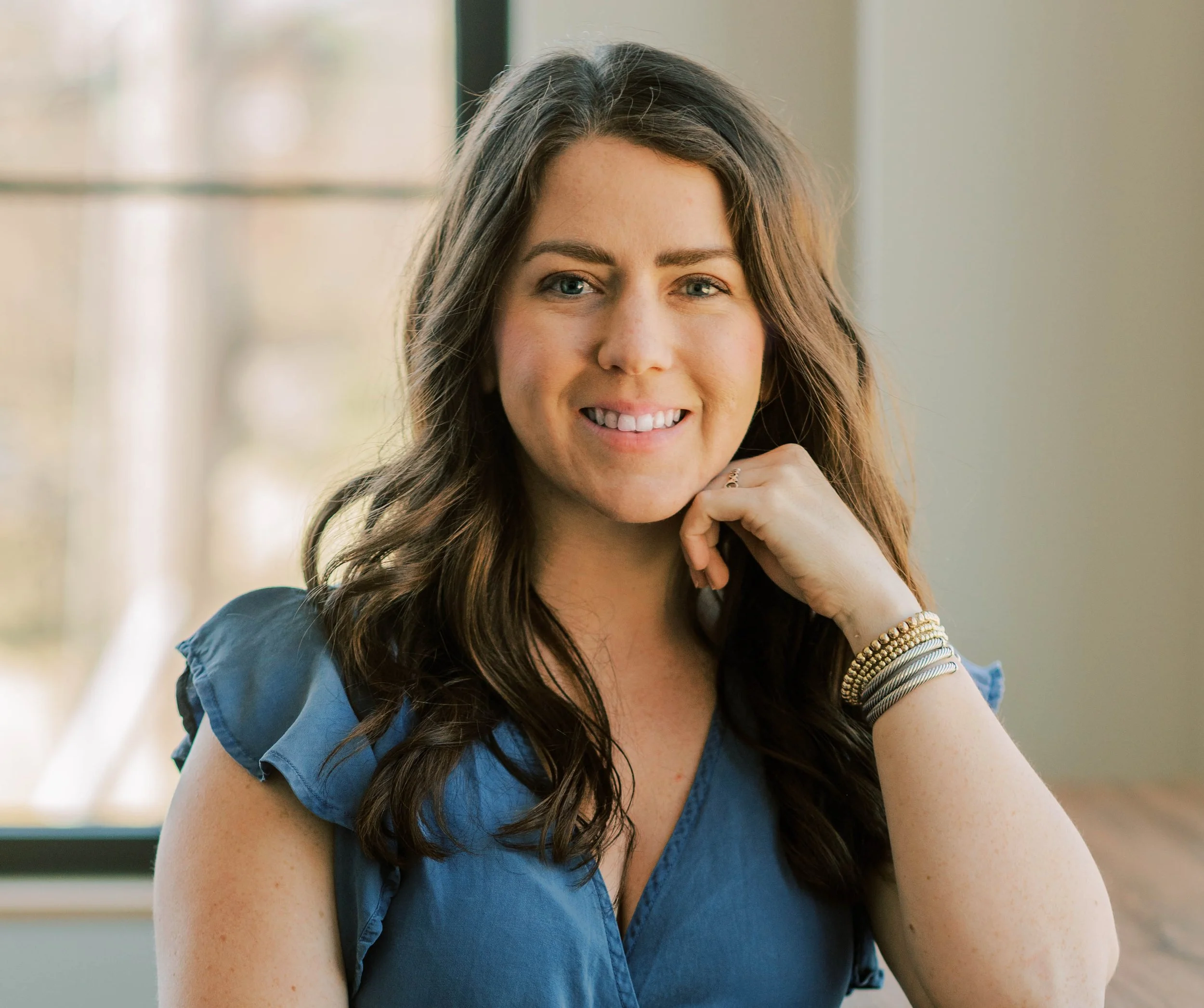 Smiling woman with long hair, wearing a blue top, seated indoors with hand resting on chin.