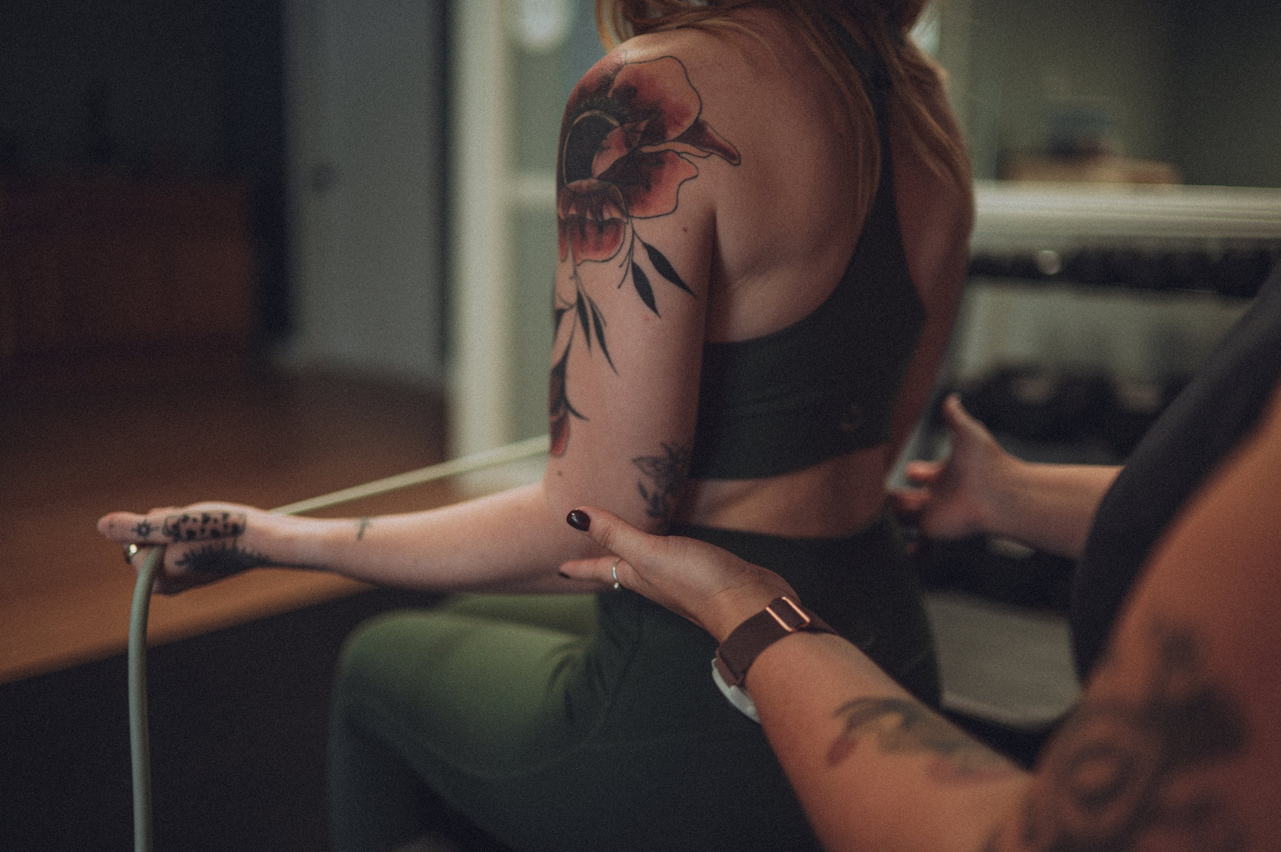 A person with tattoos on their arm and hand holding a yoga or stretching strap, with another person assisting them, inside a room with soft lighting.