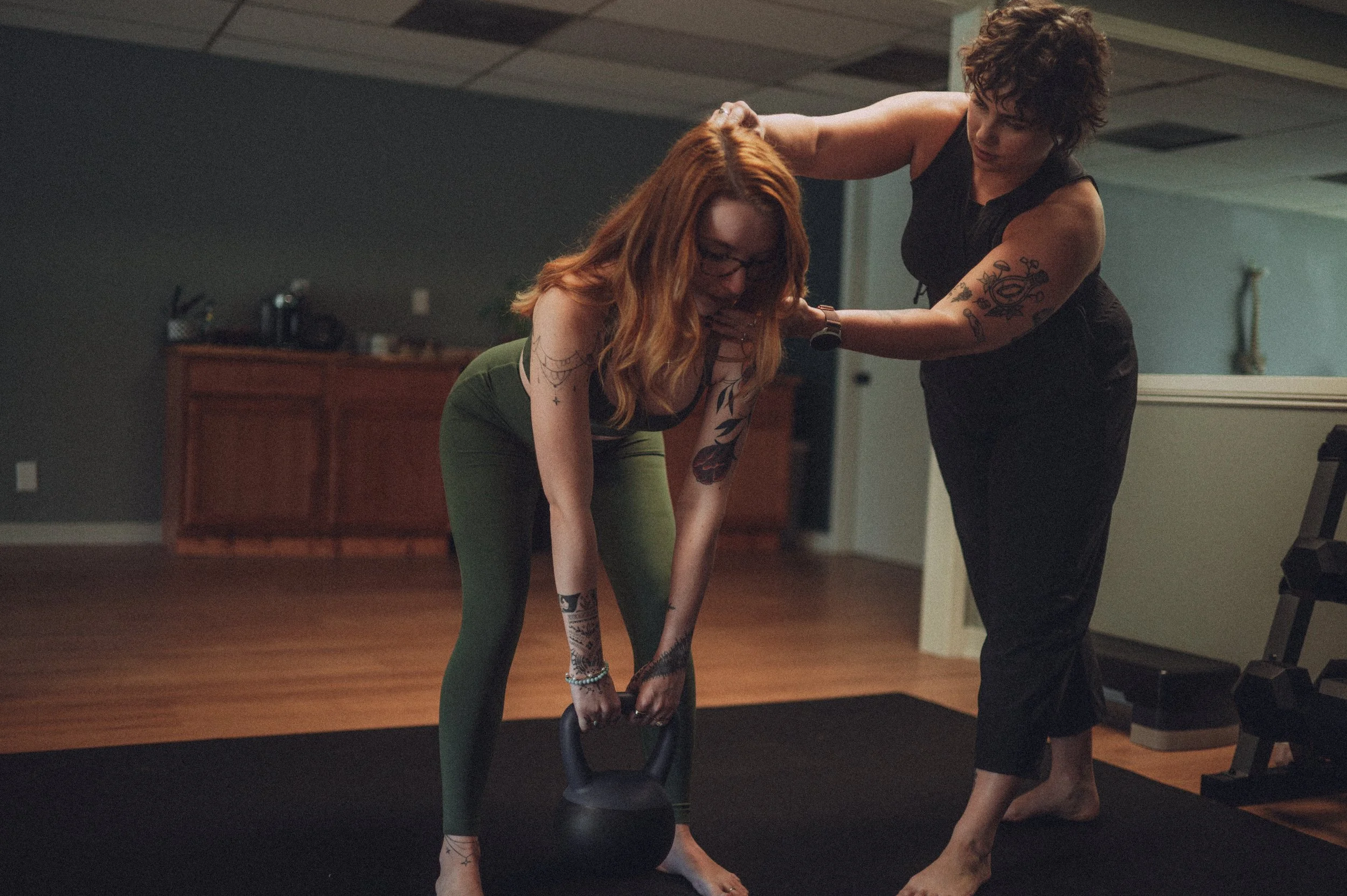 Two women in a gym, one woman is helping the other with a kettlebell exercise, both are focused on the workout. One woman has tattoos and red hair, wearing glasses and green leggings; the other has curly dark hair and is wearing a black sleeveless top.
