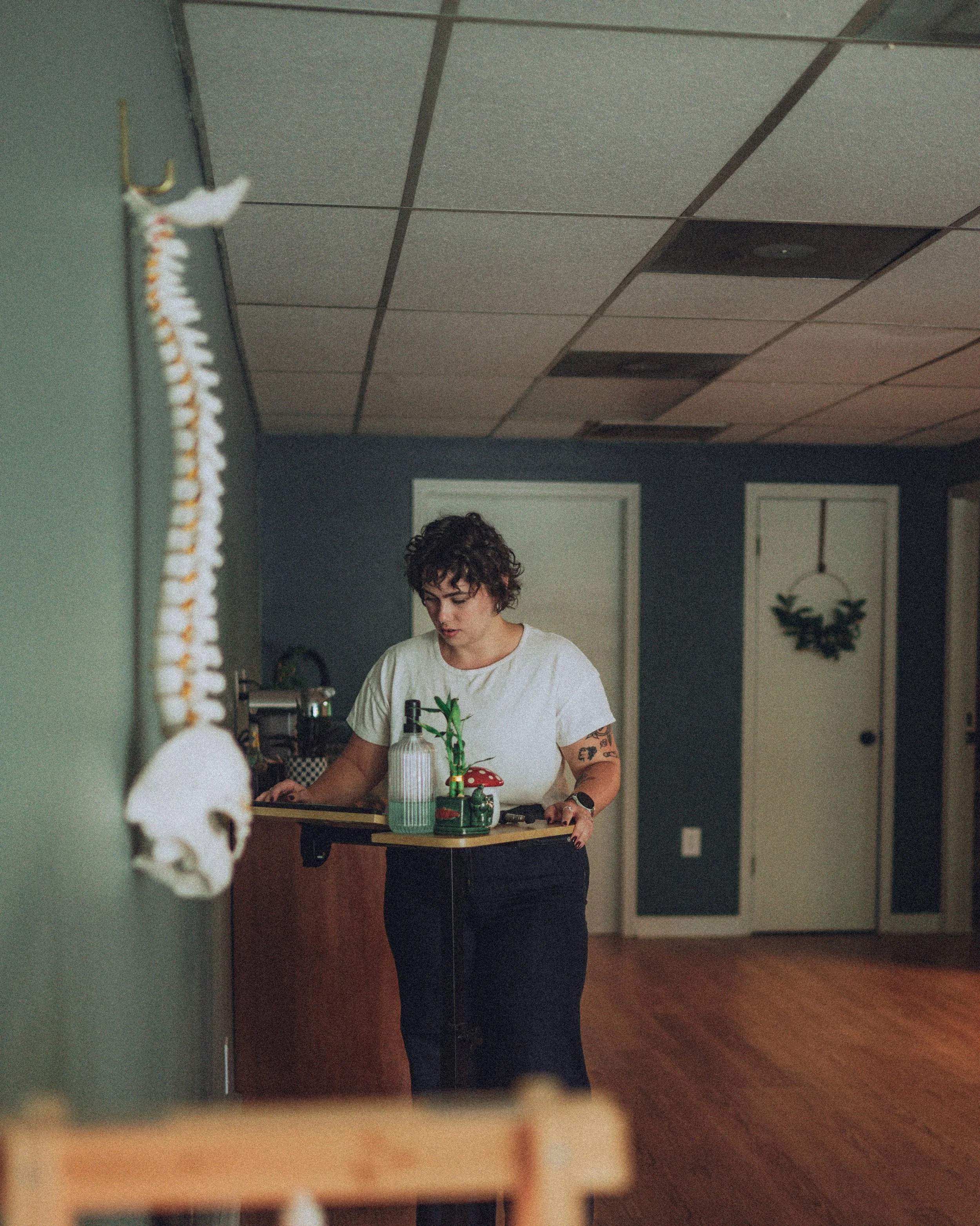 A woman with short curly dark hair is standing in a room with a wooden floor and dark walls, looking down at a table with various small decorative items, including a white vase with green plants, a red and white mushroom ornament, and a small black figurine.