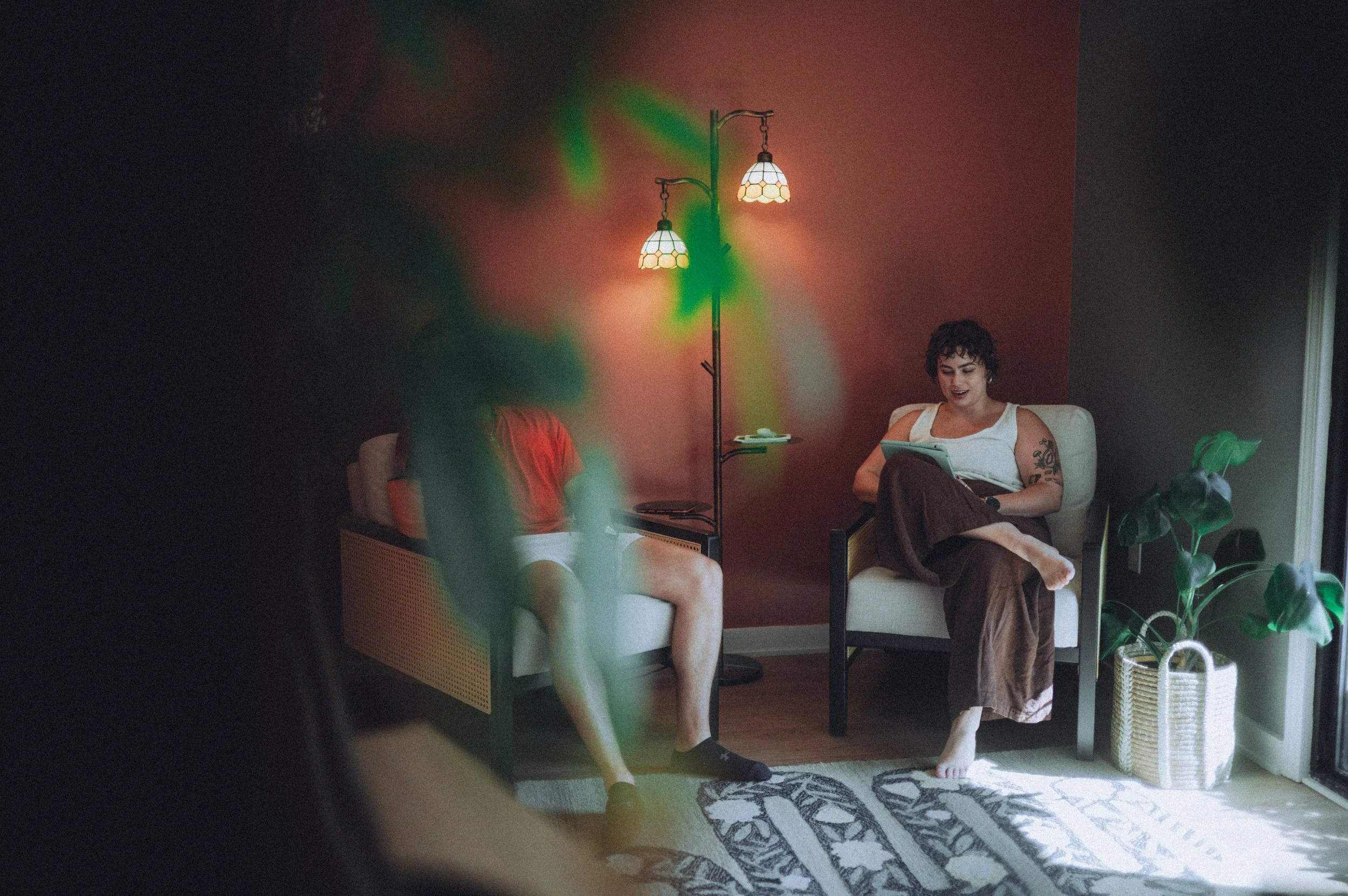 Two women sitting in chairs, one reading a tablet and the other looking down, with a vintage-style floor lamp casting warm light in a cozy room with a red wall and a potted plant nearby.