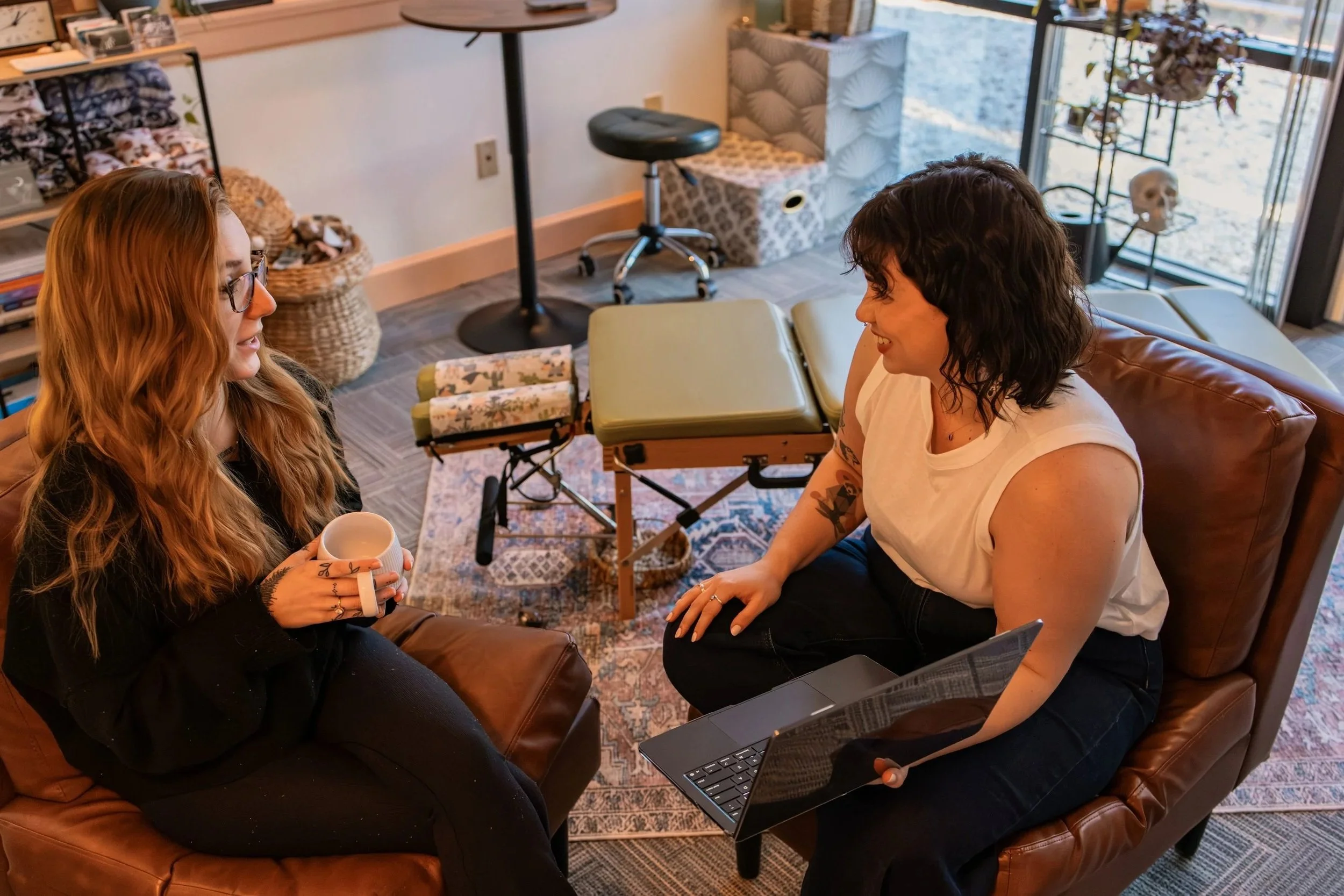 Two women sitting on sofas in a cozy room, engaging in conversation, with one holding a coffee mug and the other using a laptop, surrounded by shelves, baskets, and windows.