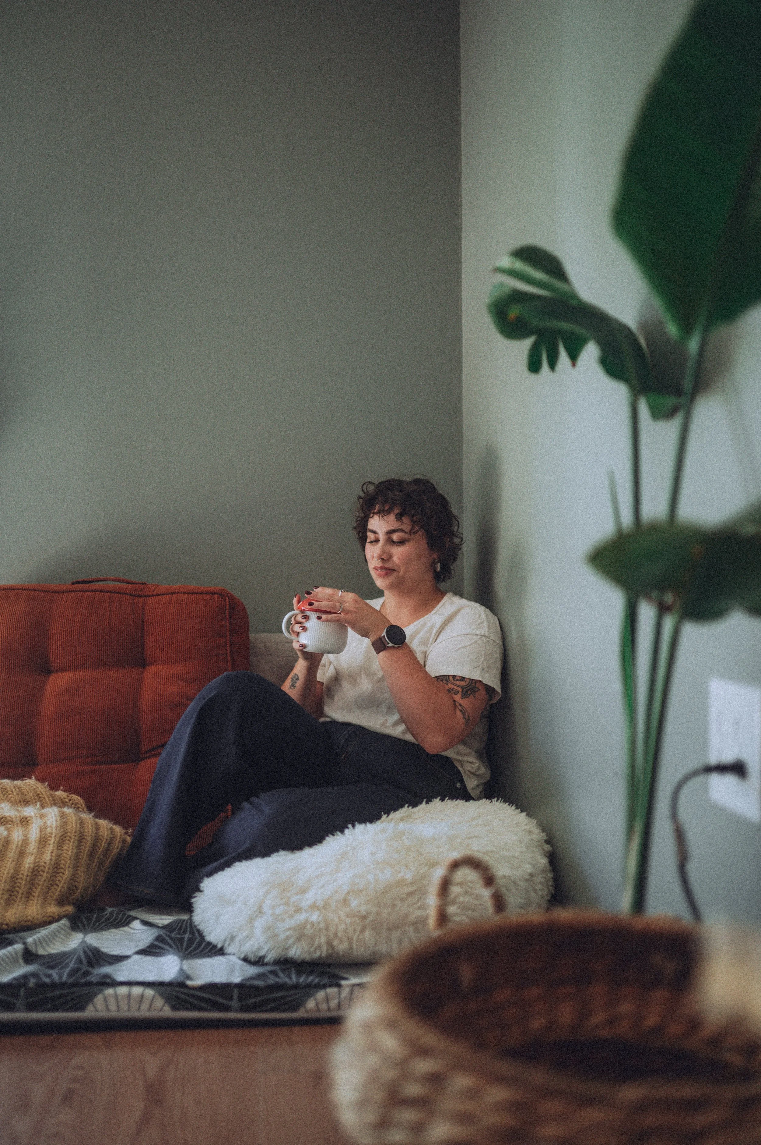 A woman with short curly hair, tattoos on her arm, and wearing a beige t-shirt, is sitting on the floor leaning against a wall. She is holding a white mug and appears to be relaxed, with a green plant partially visible on the right side of the image. There is a red couch behind her, and she is sitting on a fluffy white rug with a patterned blanket and a pair of white sneakers nearby.
