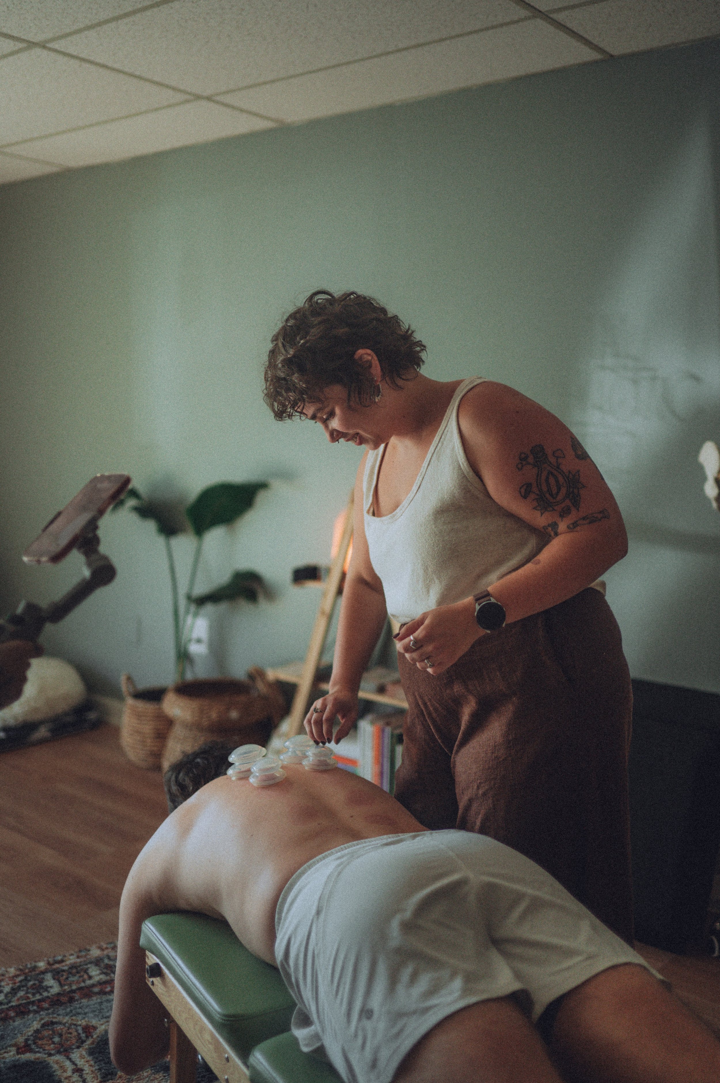A woman receiving a cup and saucer therapy treatment on her back while lying face down on a massage table. The therapist is standing beside her with a tattooed arm.