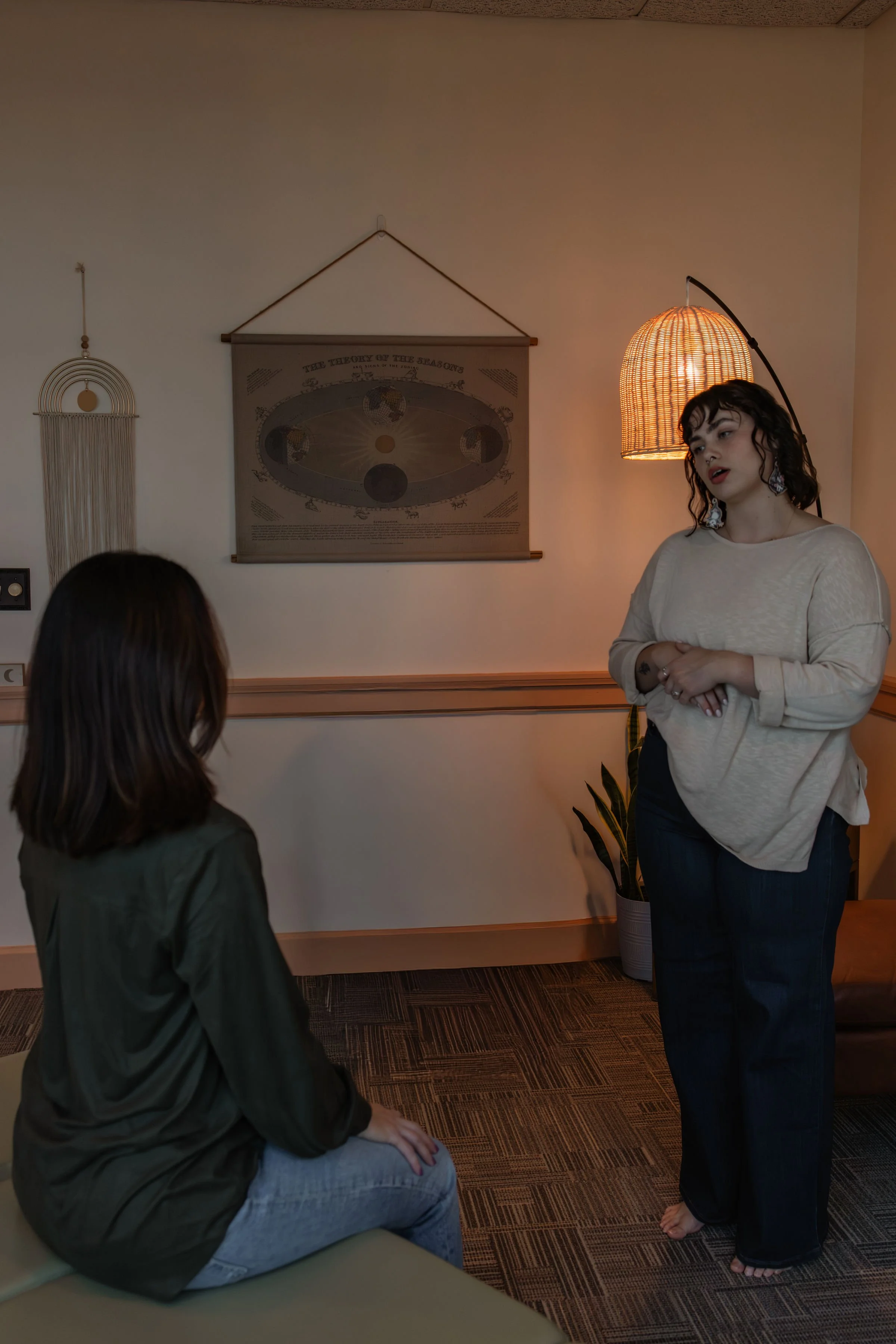 Two women engaged in conversation in a cozy room with warm lighting. One woman is sitting on a bench, facing away, wearing a dark green jacket and gray jeans. The other woman is standing, wearing a beige top and black pants, with curly hair and large earrings.