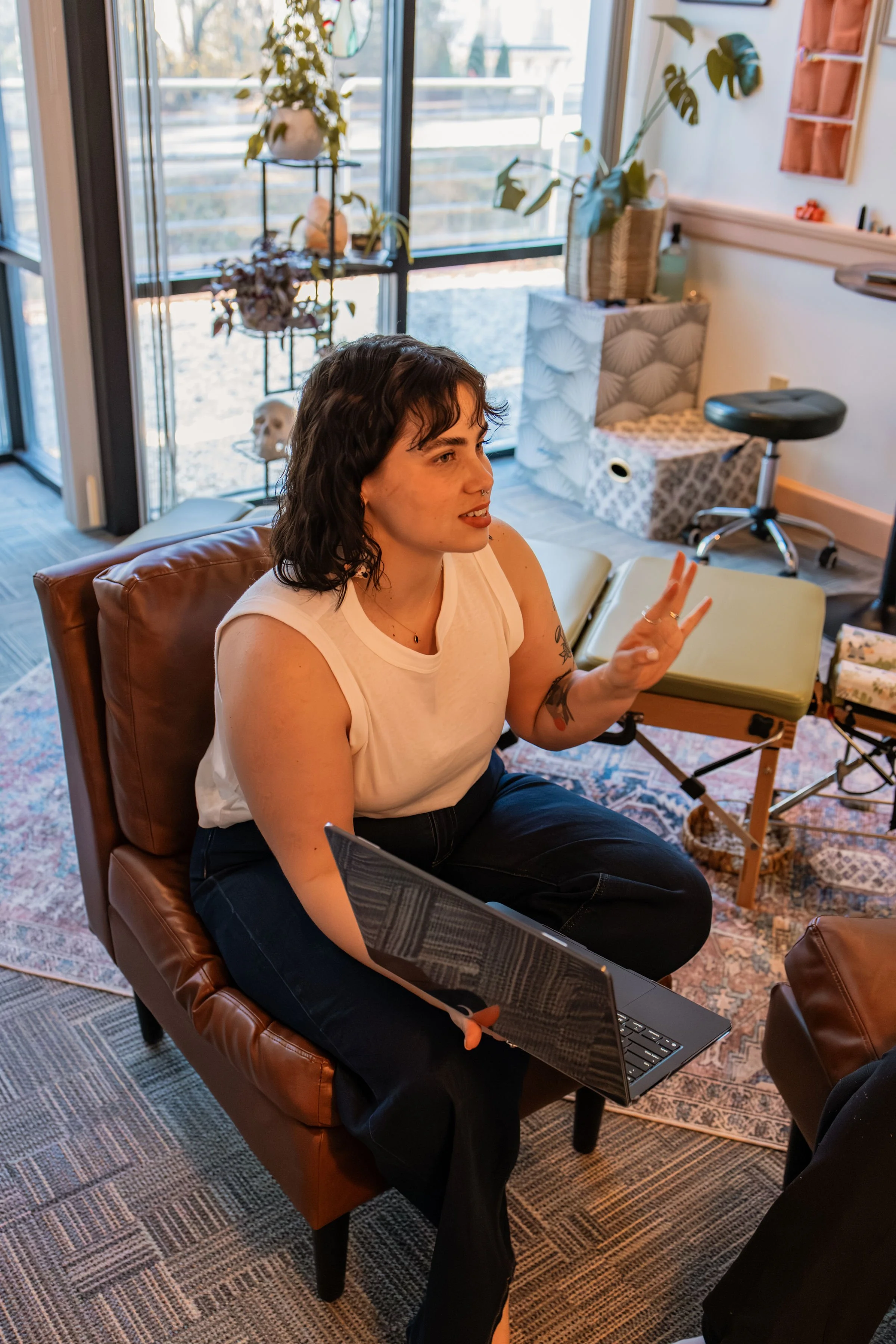 A woman with short dark hair and tattoos on her arms holding a laptop, sitting on a leather chair and gesturing with her hand during a conversation in a cozy, well-lit room with plants, a window, and various decorative items.