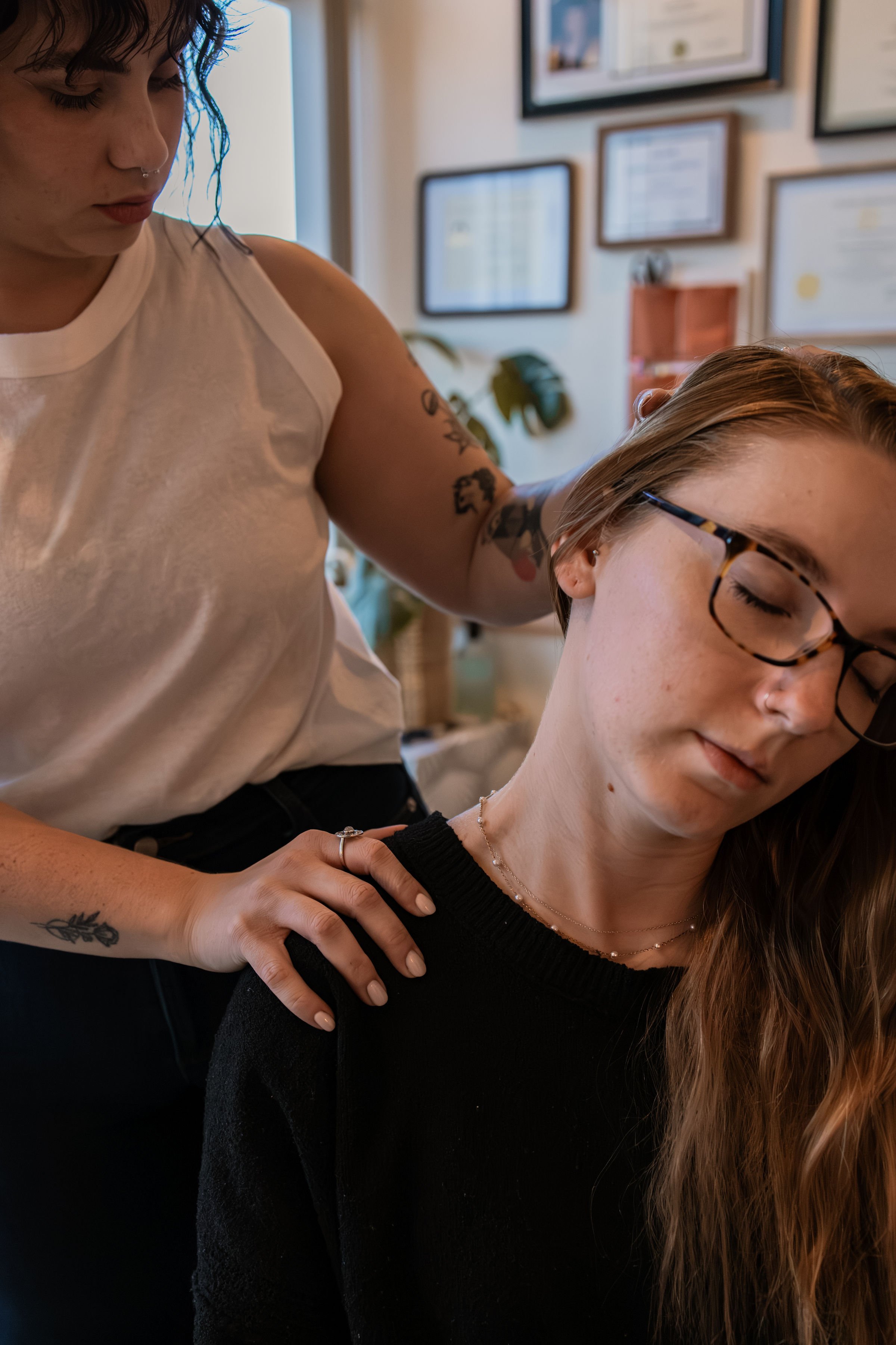A woman with short brown hair, glasses, and tattoos on her arms is giving a shoulder stretch to a young woman with long brown hair and glasses, who is sitting with her eyes closed in a cozy room decorated with framed certificates and plants.