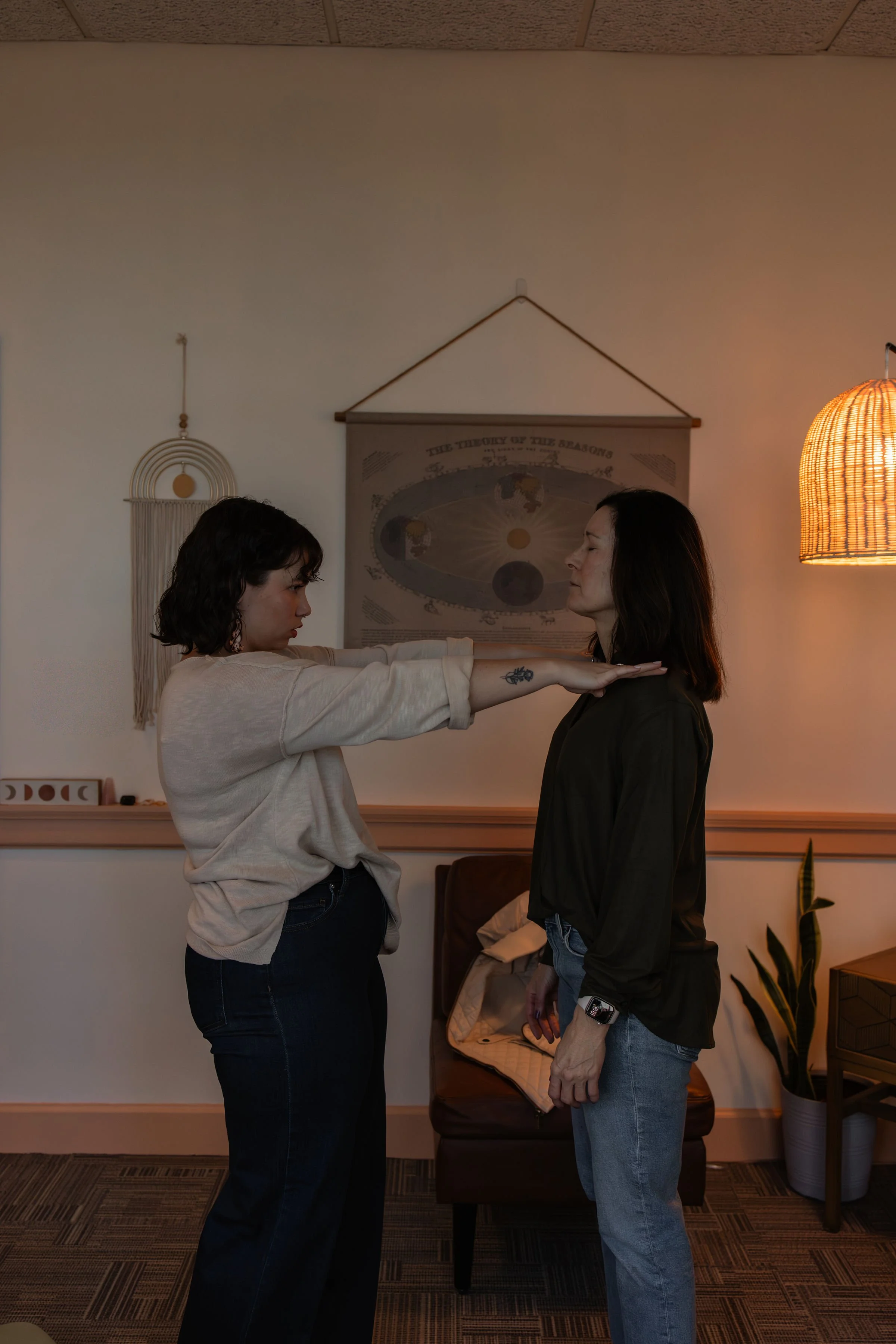 Two women in a dimly lit room, standing face to face, with one woman extending her arms horizontally, palms down, toward the other woman. The room has wall decor including a circular wall hanging and a framed chart, a chair, a plant, and a warm-toned hanging lamp.