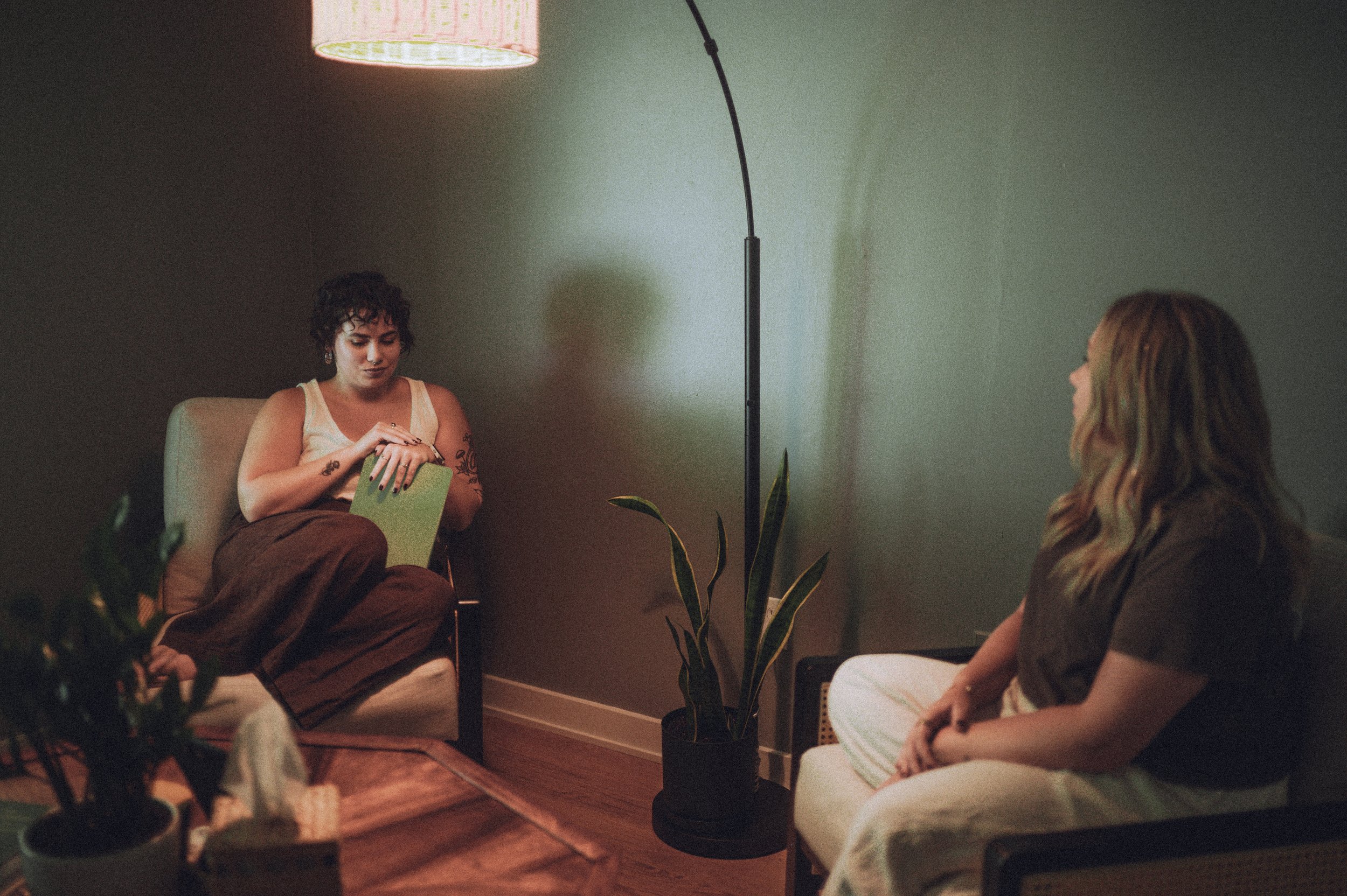 Two women sitting in a dimly lit room, engaging in conversation. One woman with dark, curly hair, tattoos, wearing a tank top, is sitting on a chair with her hands on her lap holding a green notebook. The other with long, wavy hair is sitting on a white chair, seen from the back. There is a tall floor lamp, a potted plant, and a green wall in the room.
