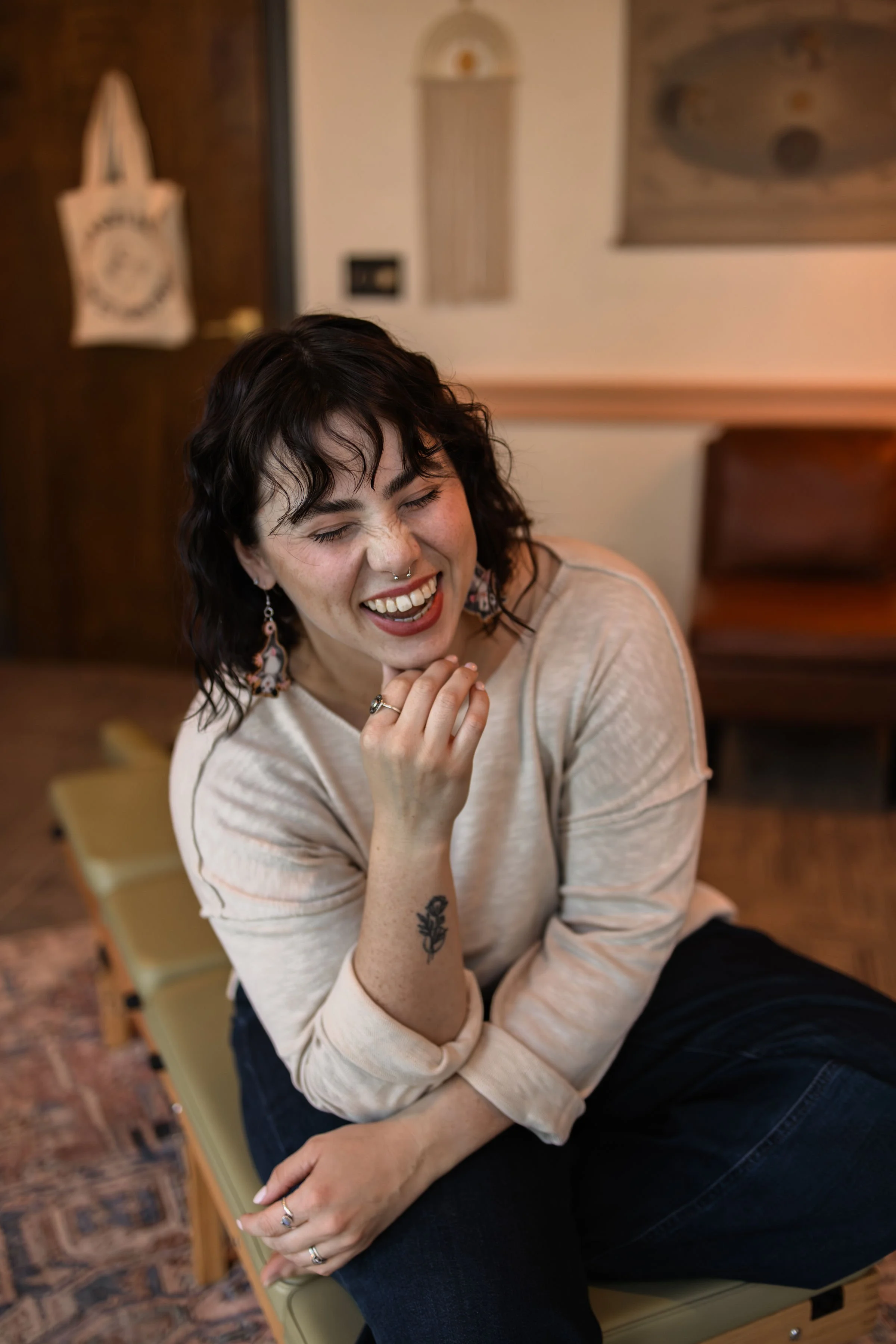 A woman with short black hair, tattoos, and earrings, laughing and smiling while sitting on a green bench in a cozy indoor space.