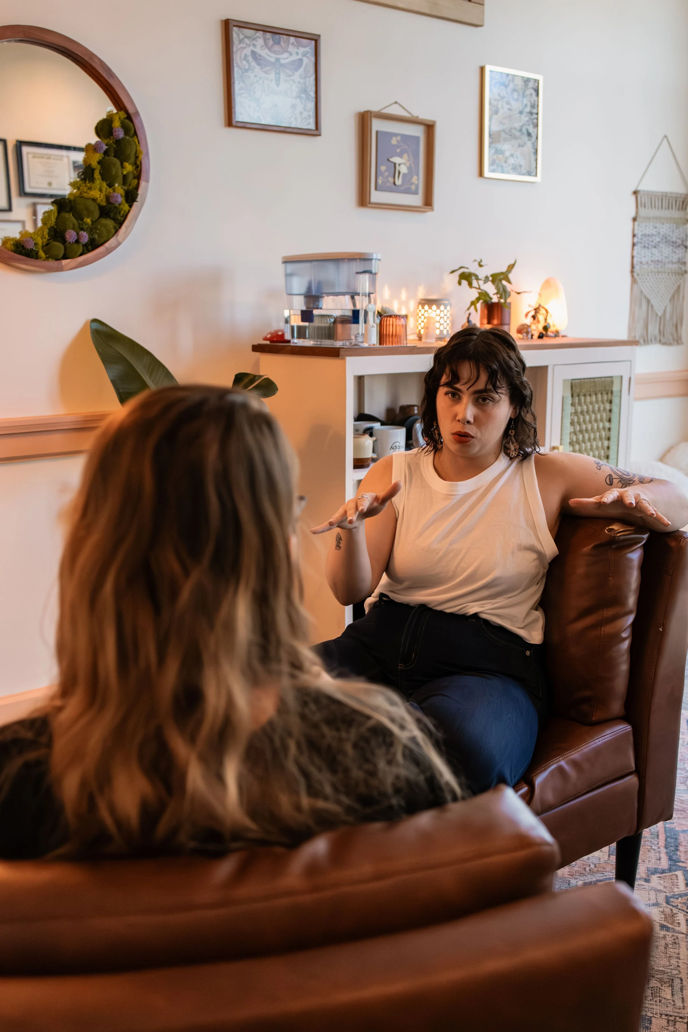 Two women having a conversation in a cozy living room with framed artwork, plants, and warm lighting.