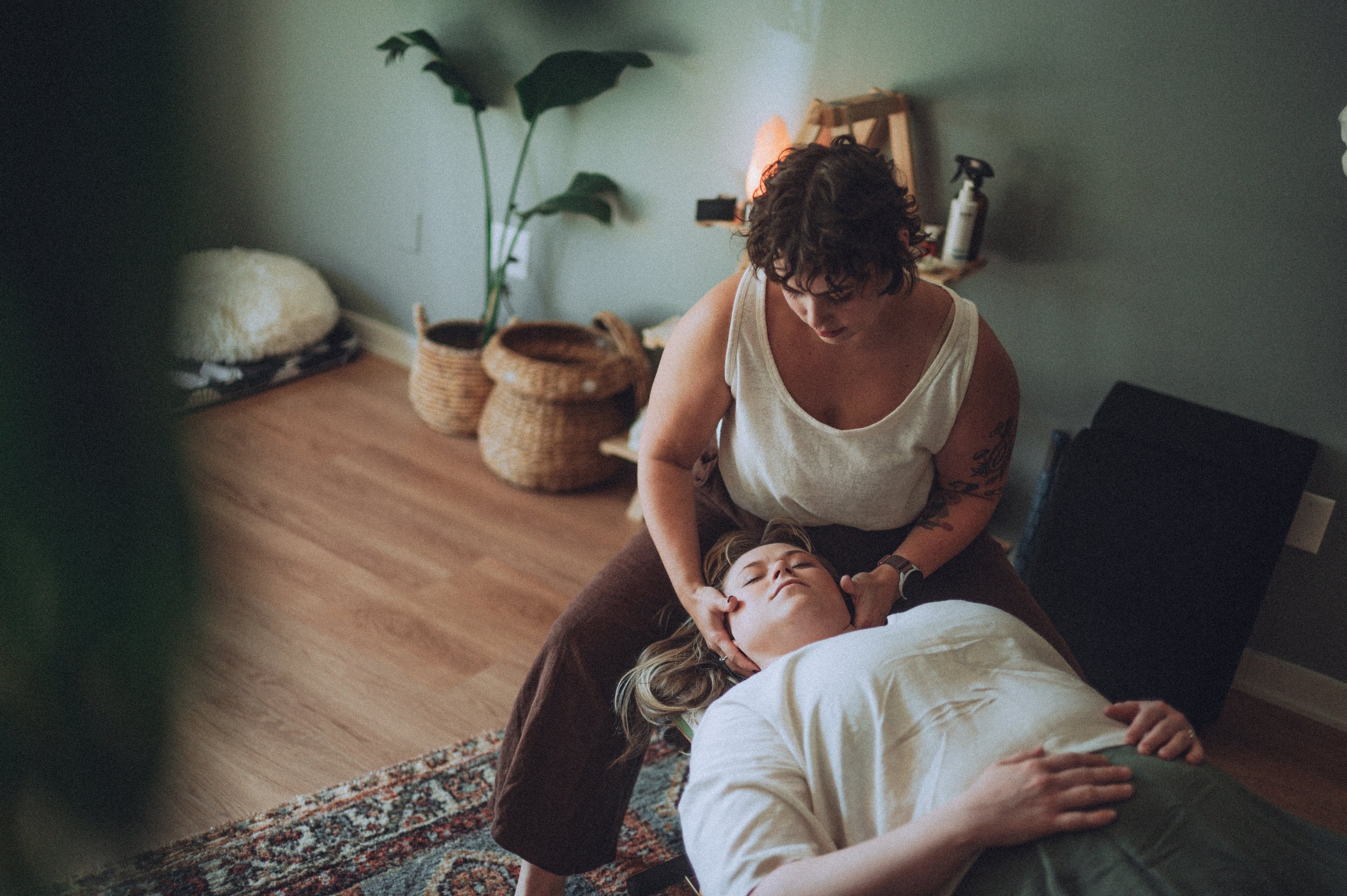 A woman giving an adjustment to a woman lying on a massage table in a cozy room decorated with baskets, plants, and a rug.