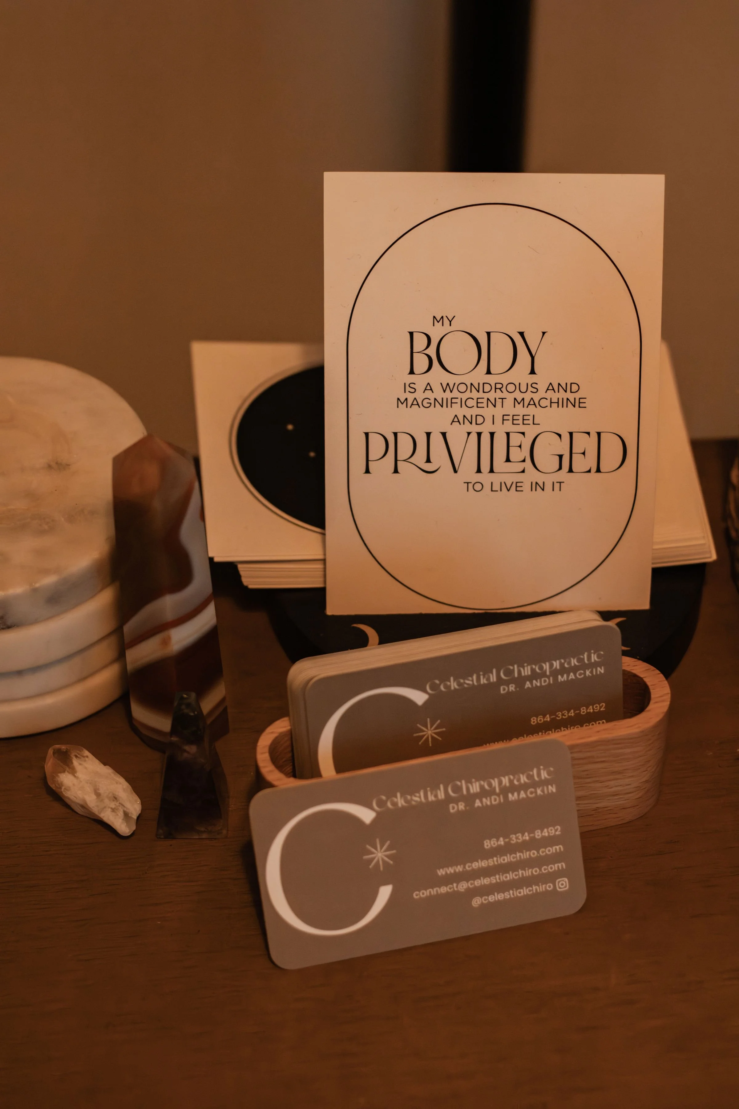 Business card display, a small wooden tray holding cards for Celestial Chiropractic, a sign with an inspirational quote about the body, and some decorative stones and mineral pieces.