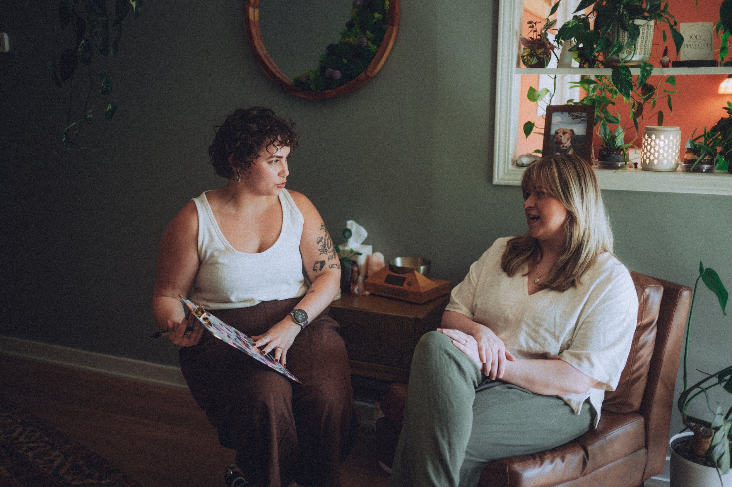 Two women sitting and conversing in a cozy, plant-filled room, with one holding a magazine and the other smiling. There is a window with decorative items and green plants on the windowsill and surrounding shelves.