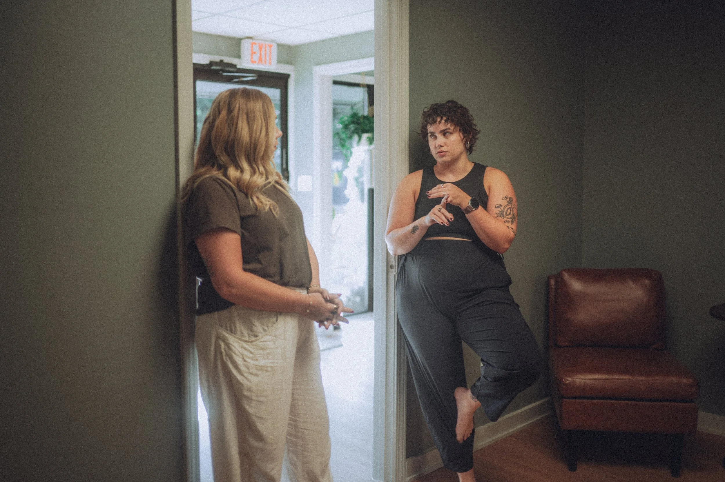 Two women are having a conversation in a hallway, one with blonde hair wearing a brown top, and the other with short curly hair wearing a black sleeveless top and black pants, leaning against the wall.