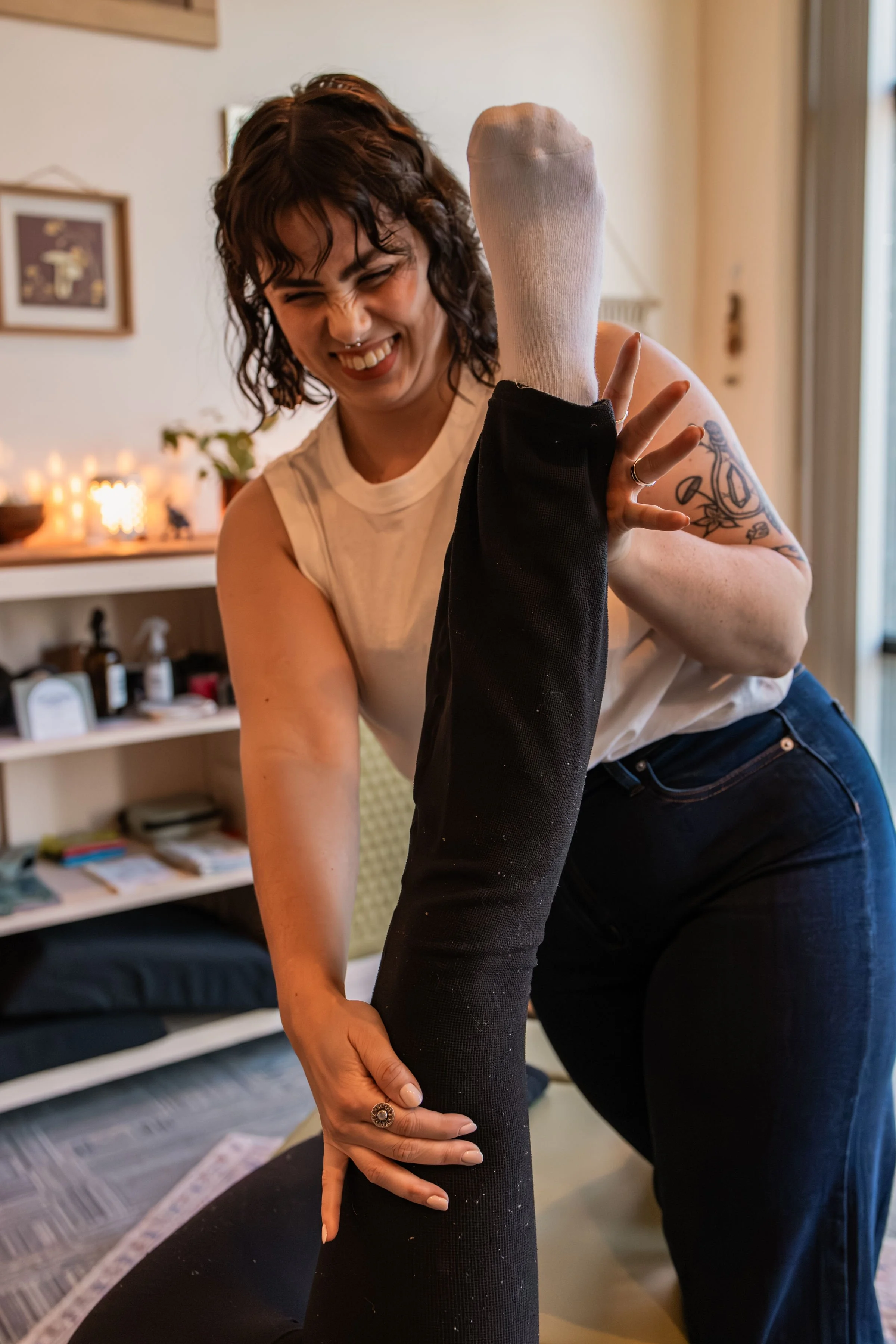 A woman with short curly hair and a nose piercing, smiling and laughing, giving a leg stretch to a person lying down with their leg raised. The room has warm lighting and decor, including candles and framed artwork.