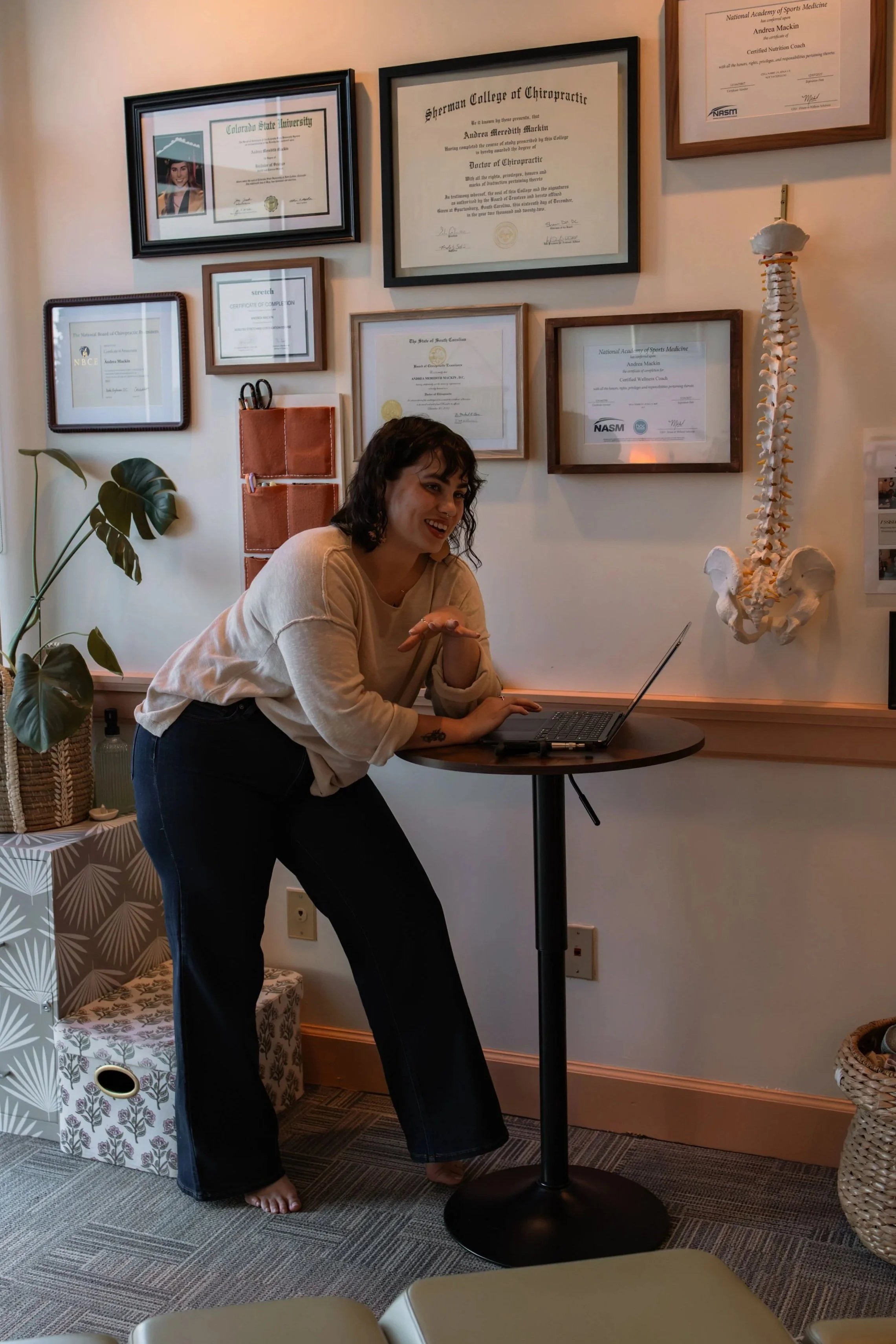 A woman with shoulder-length dark hair, smiling and leaning on a small round table with a laptop, in front of a wall decorated with framed certificates and diplomas, a potted plant, and a model of a human spine.