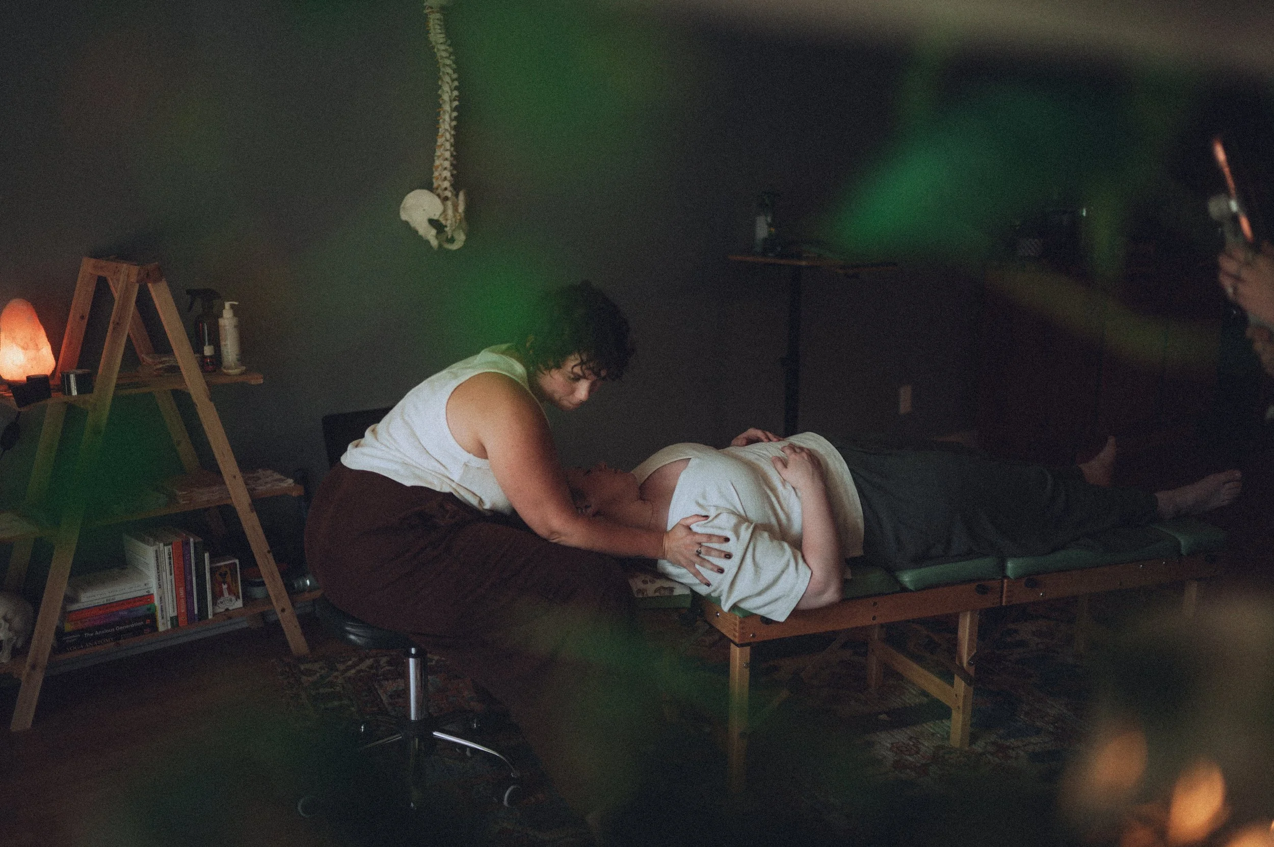 A woman performs a Reiki healing session on a woman lying on a massage table in a dimly lit room, with candles and books in the background.
