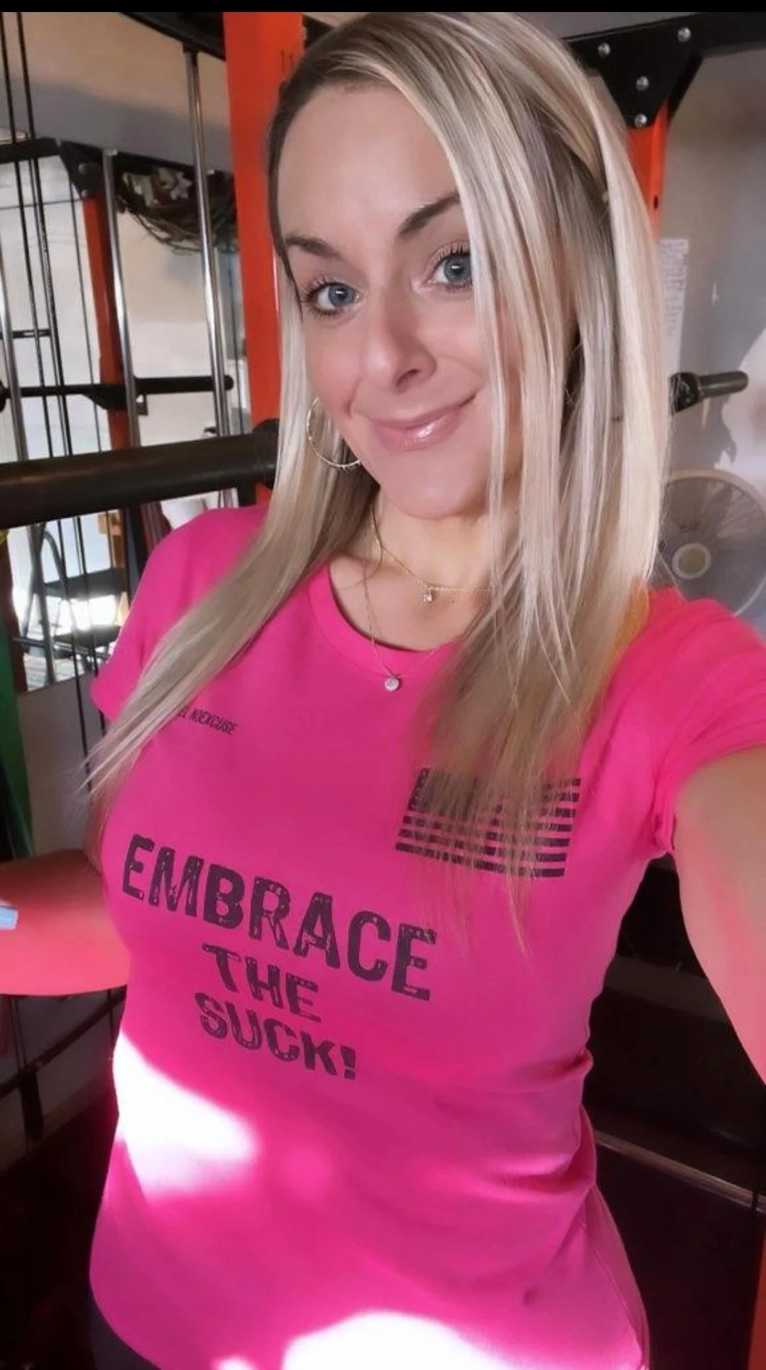 Woman in a gym wearing a pink shirt with "Embrace the Suck!" printed on it.